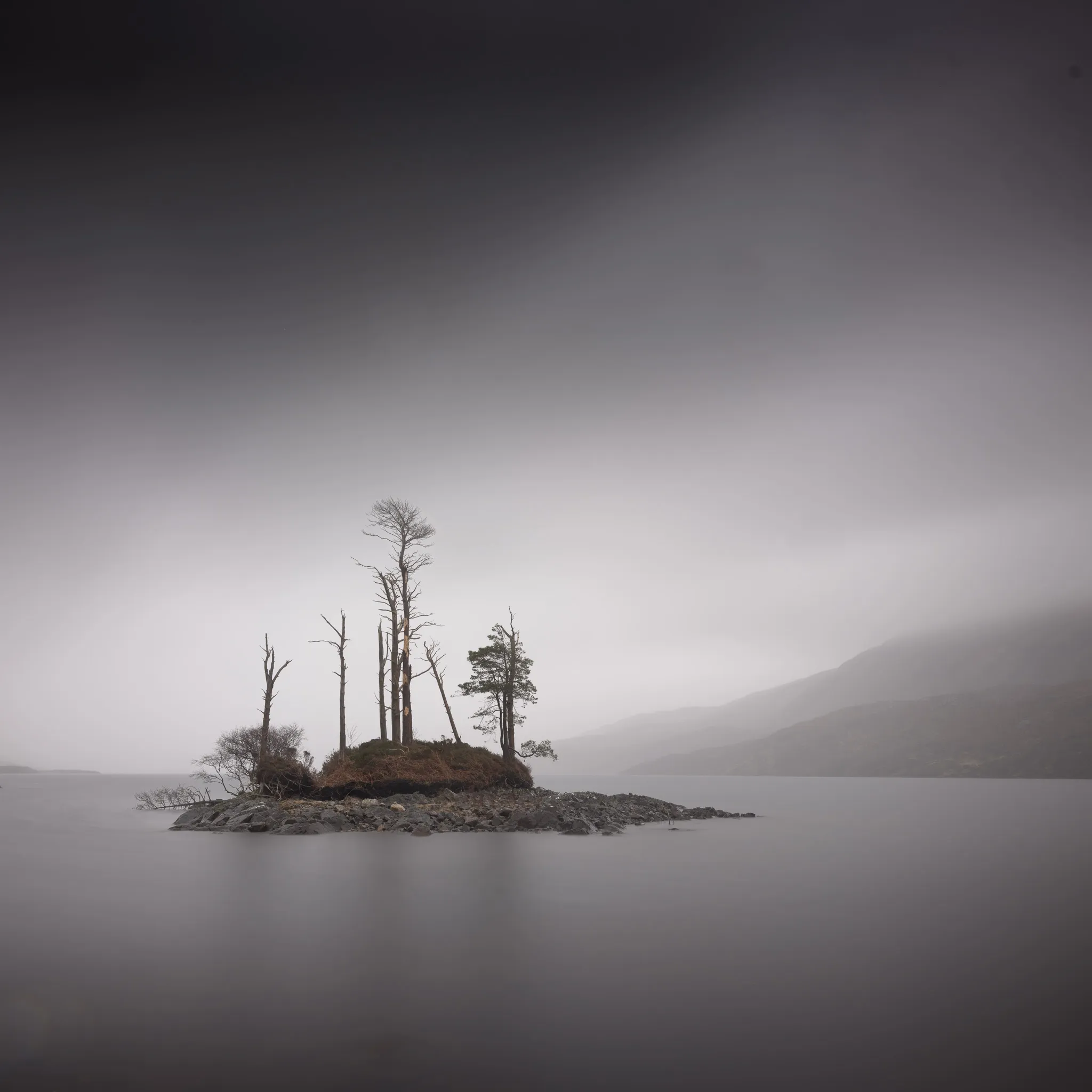A small rocky island crowned with dead and living pines on a still, misty loch, a mountain fading into fog beyond