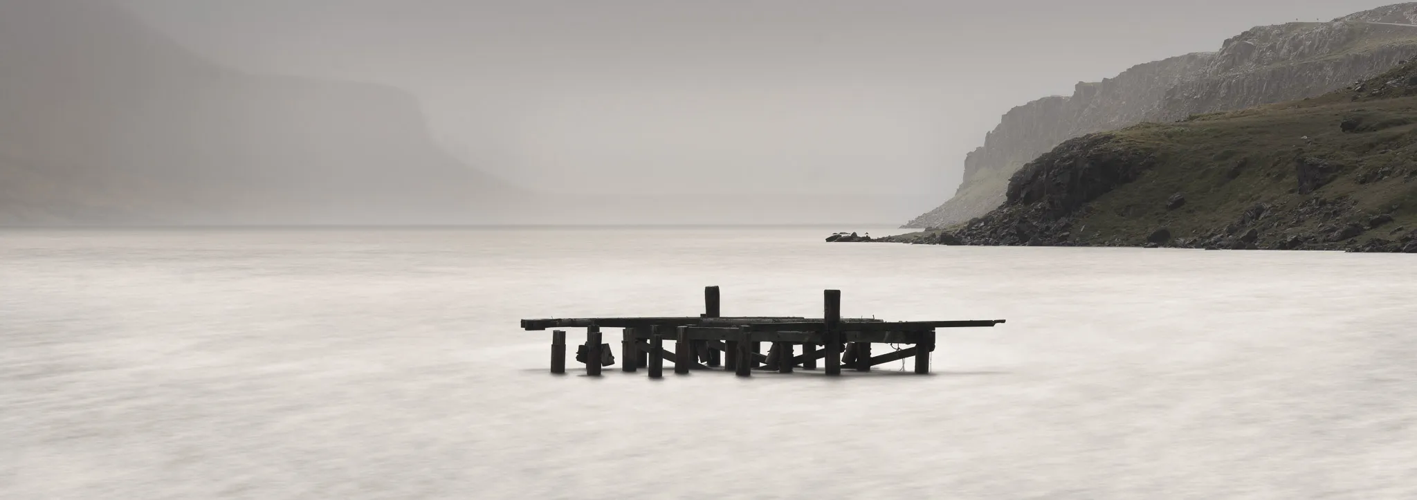 The skeletal remains of an old wooden jetty in smooth, misty water below fog-shrouded cliffs