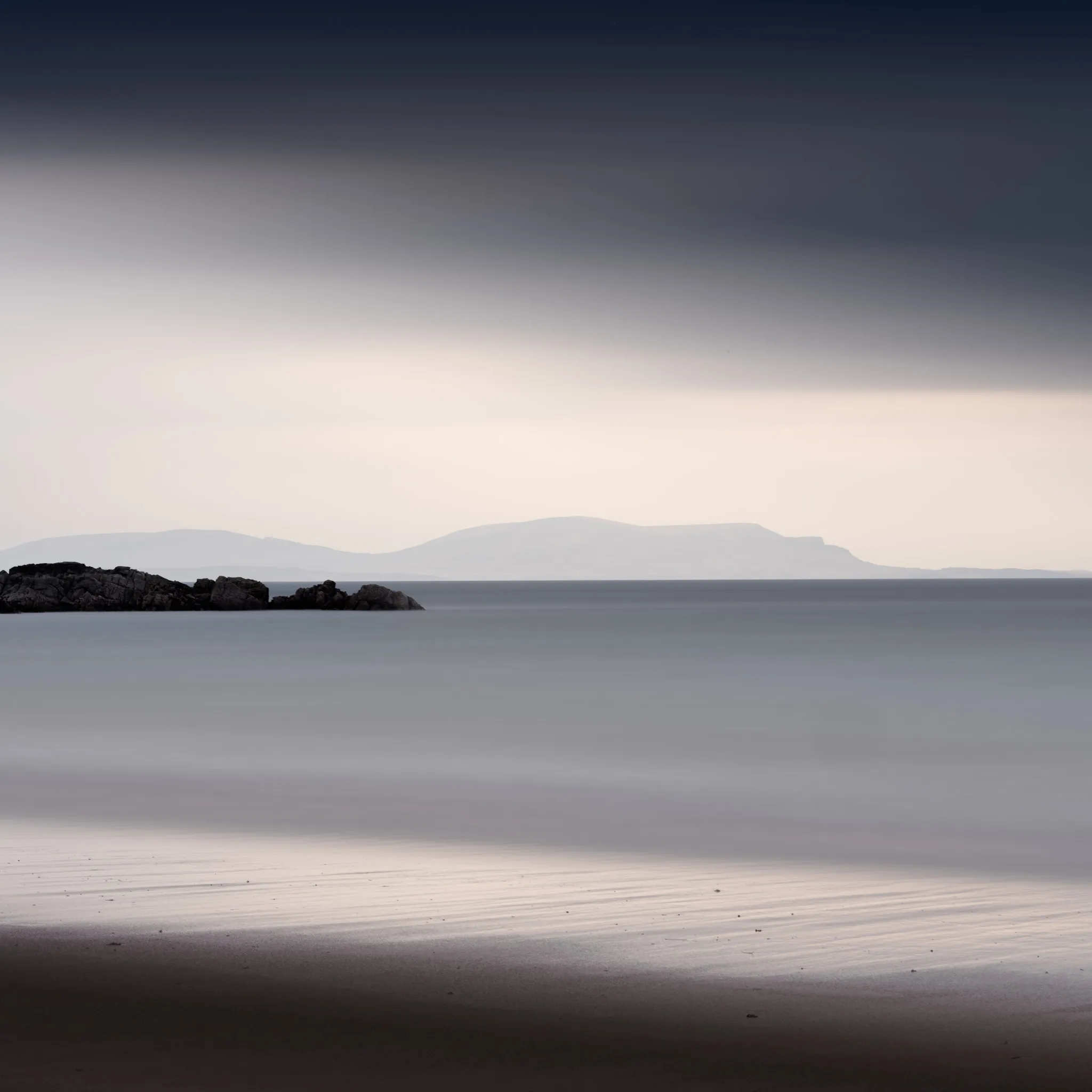 A low rocky outcrop on a calm beach at dusk, flat-topped hills silhouetted across still water