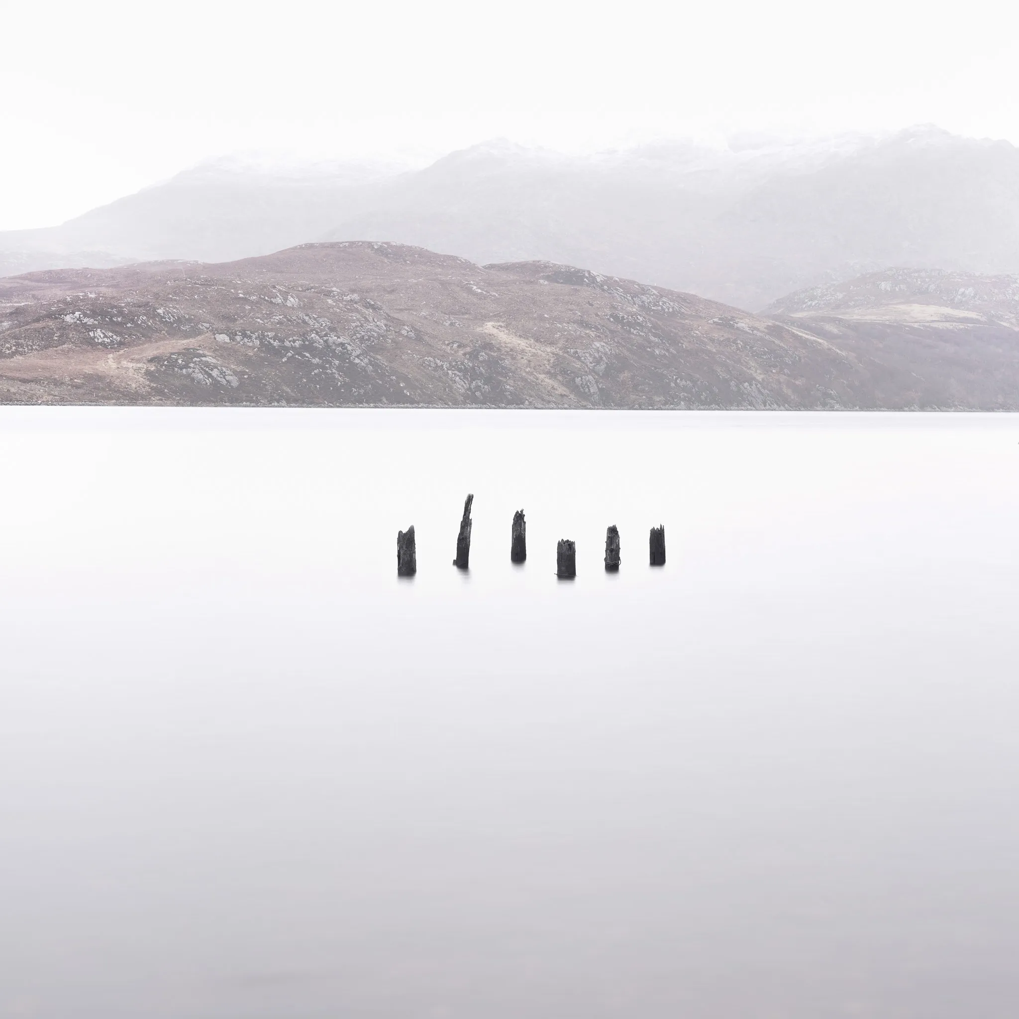 Six weathered wooden posts standing in a mist-smooth loch below bare hills