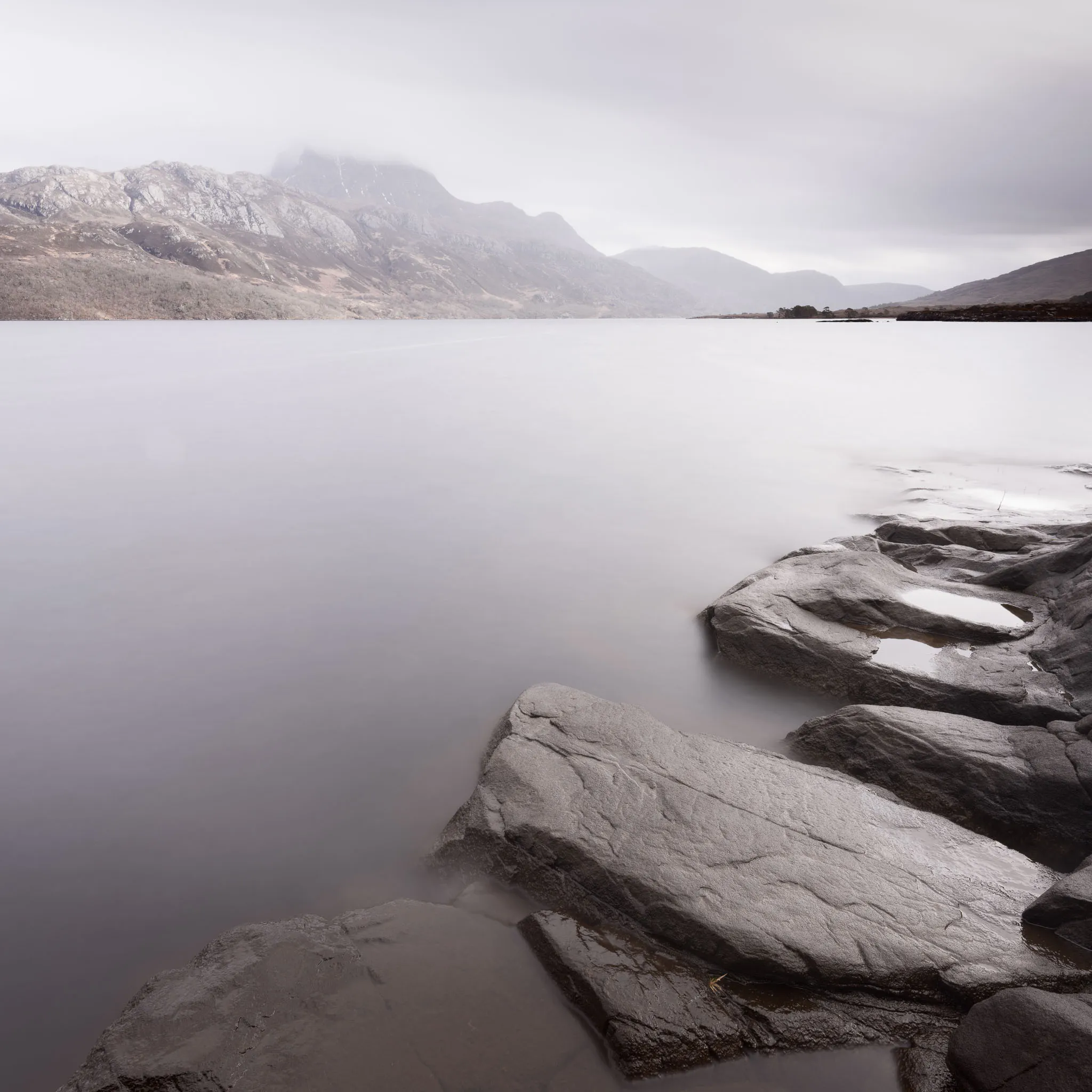 Wet foreground rocks on a still loch beneath a snow-streaked mountain wreathed in cloud