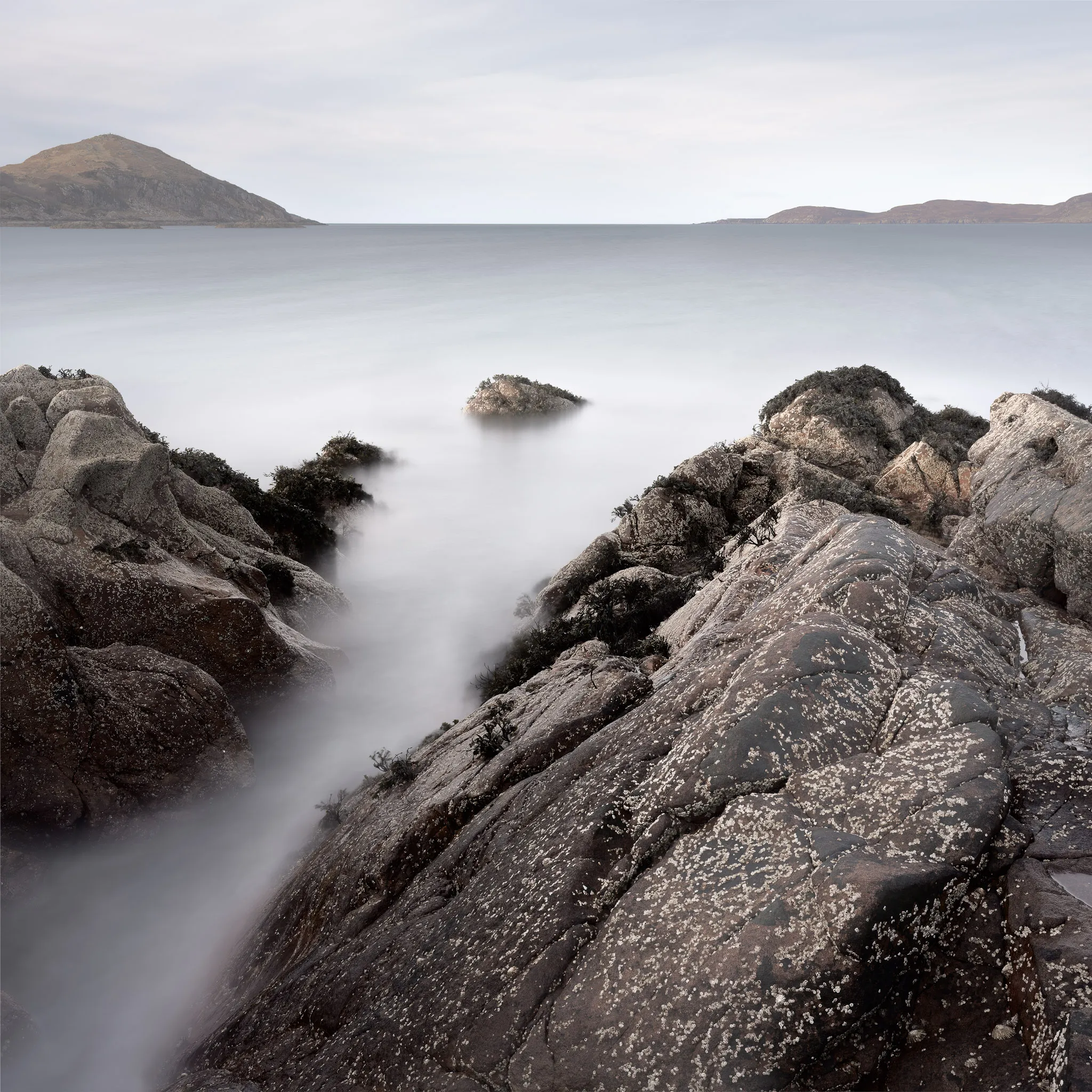 Lichen-flecked coastal rocks flanking a misty inlet, low hills across a calm sea