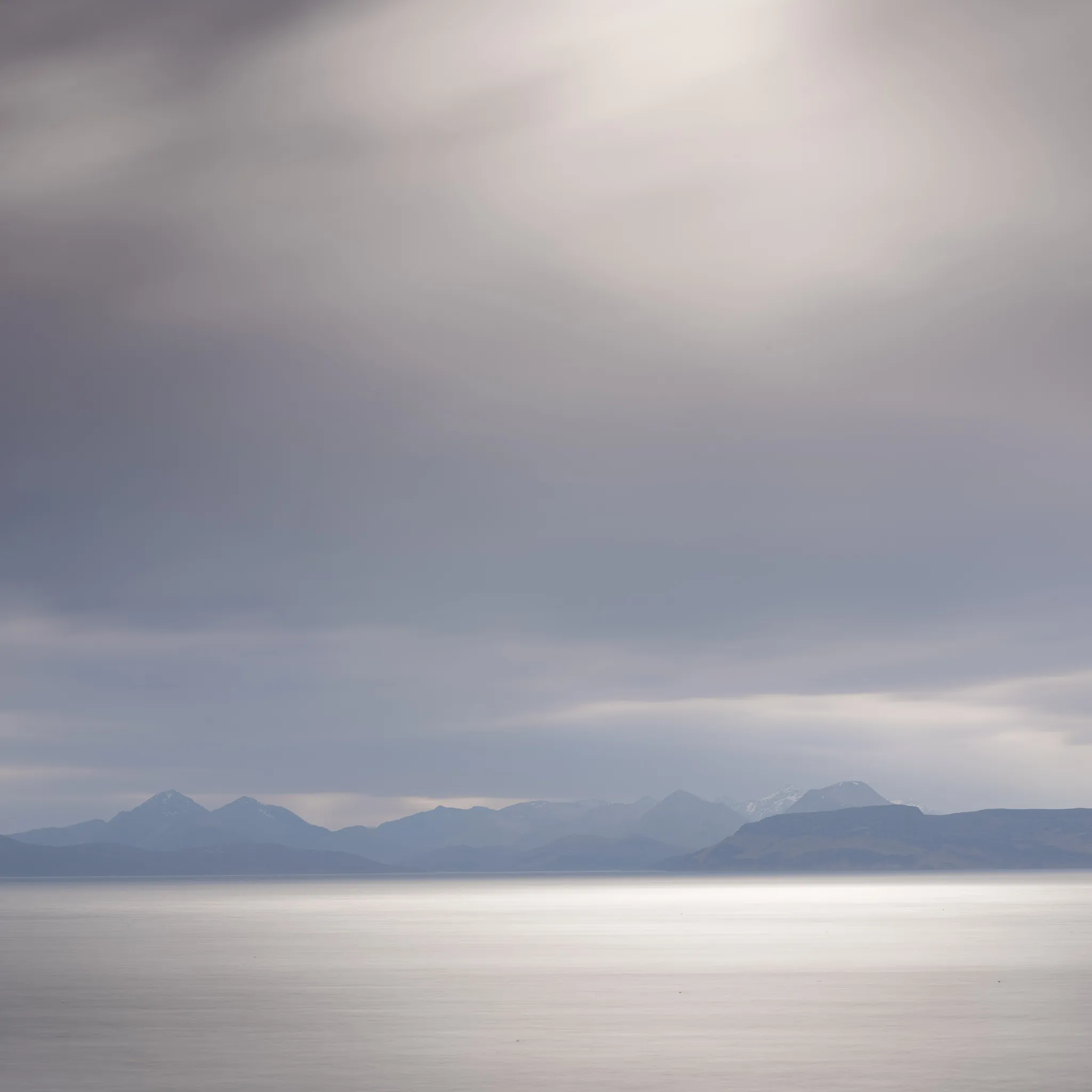 A silky calm sea below a distant silhouetted mountain range under a vast soft grey sky