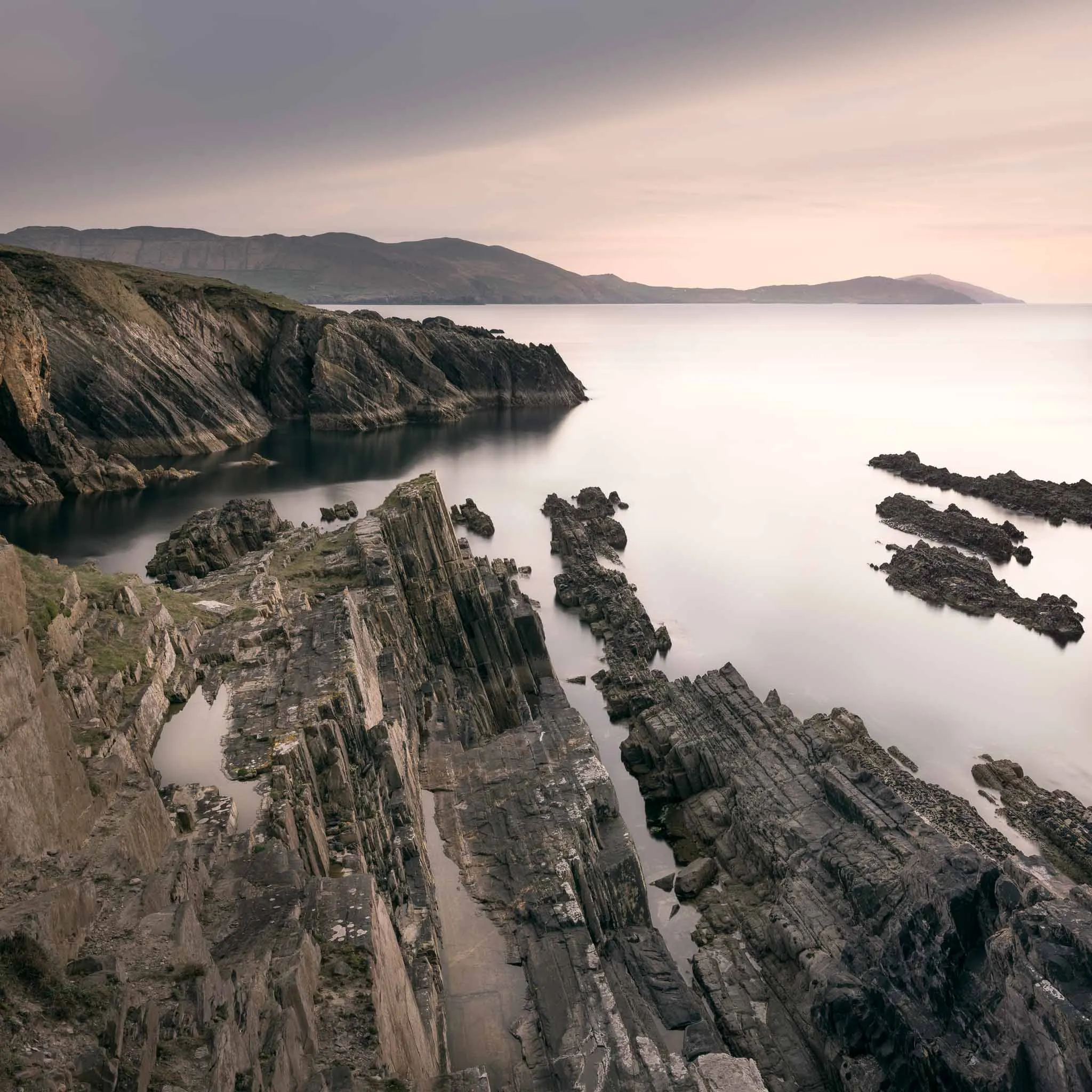 Tilted layers of dark slate rock along the coast at dusk, calm sea and a headland under a peach sky