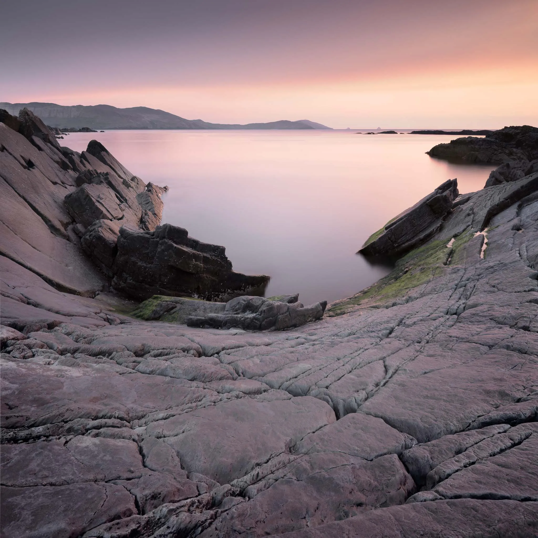 Angular rock strata framing a smooth, lilac-toned sea at sunset with distant headlands
