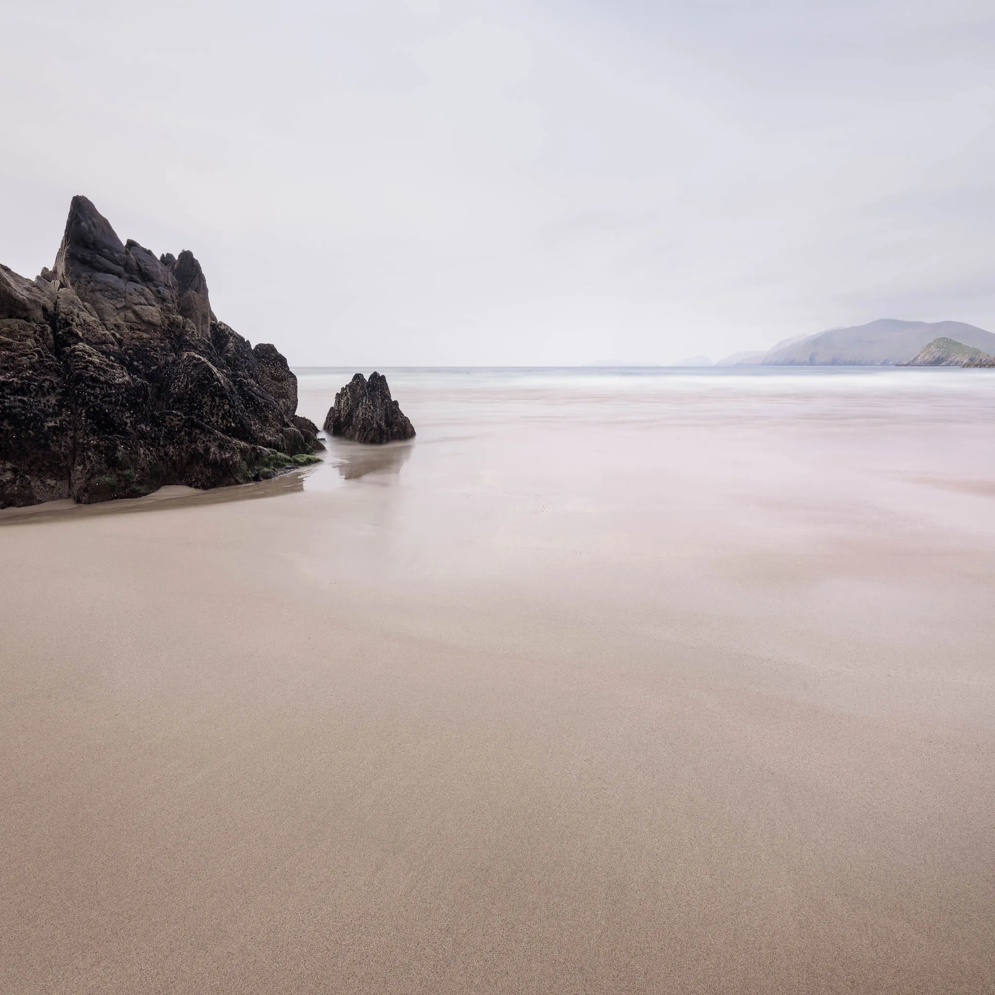 A wide, empty sandy beach with a jagged rock outcrop and a calm pastel sea, distant headland