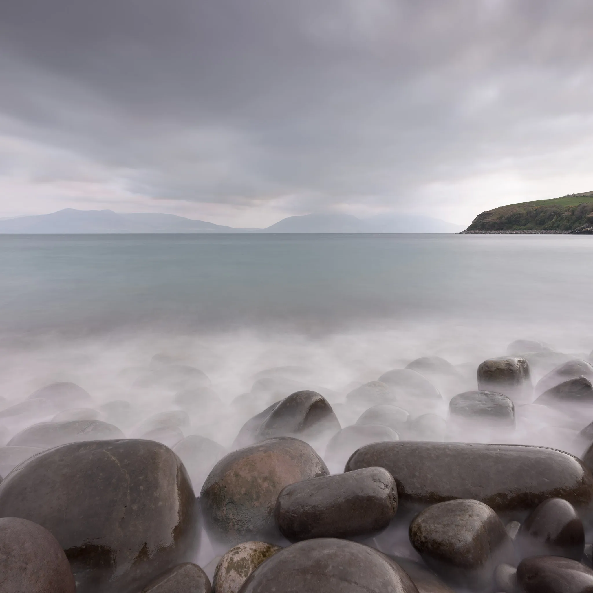 Rounded wet boulders and long-exposure surf on the shore, mountains across the bay under a grey sky