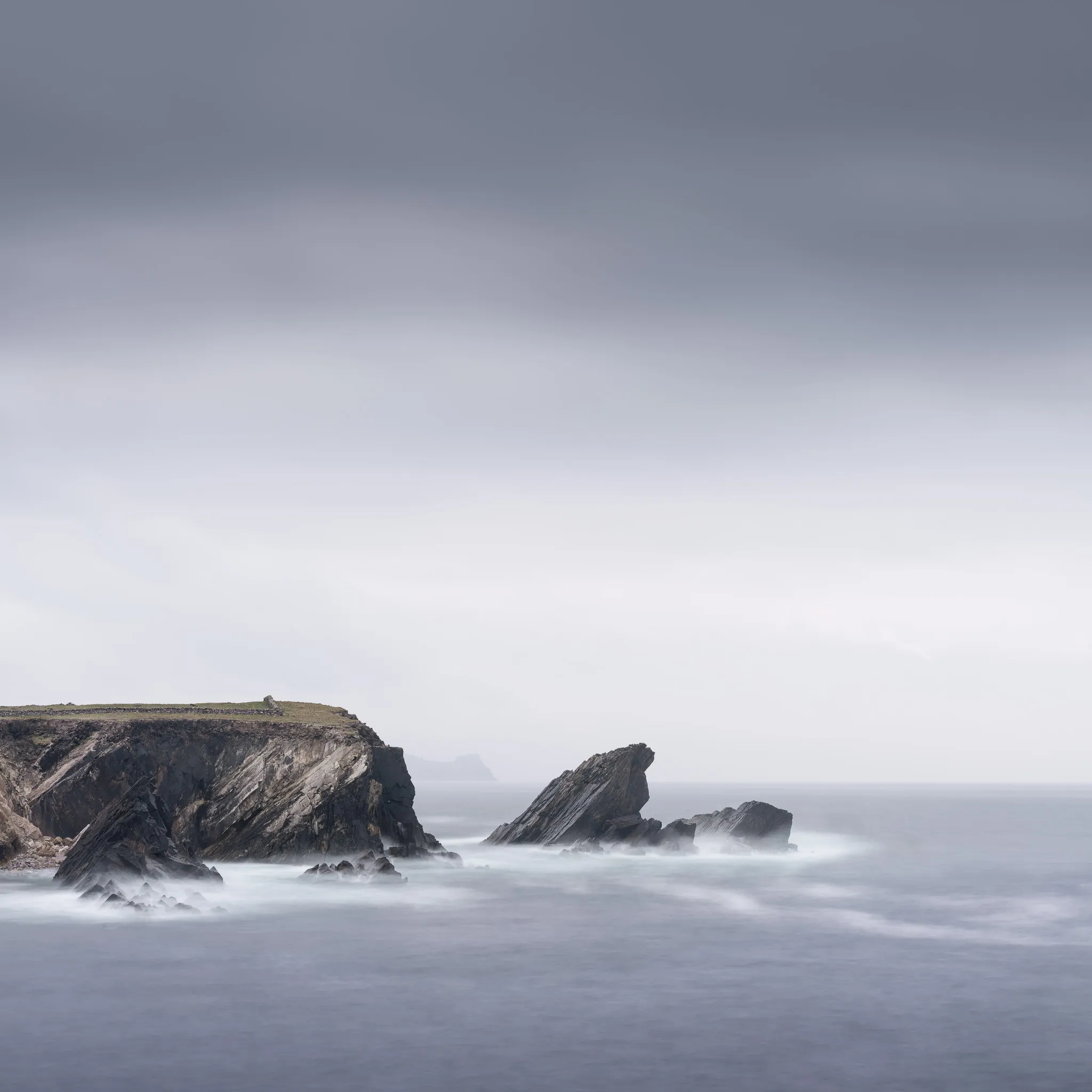A rugged cliff headland and jagged rocks amid long-exposure surf under an overcast sky