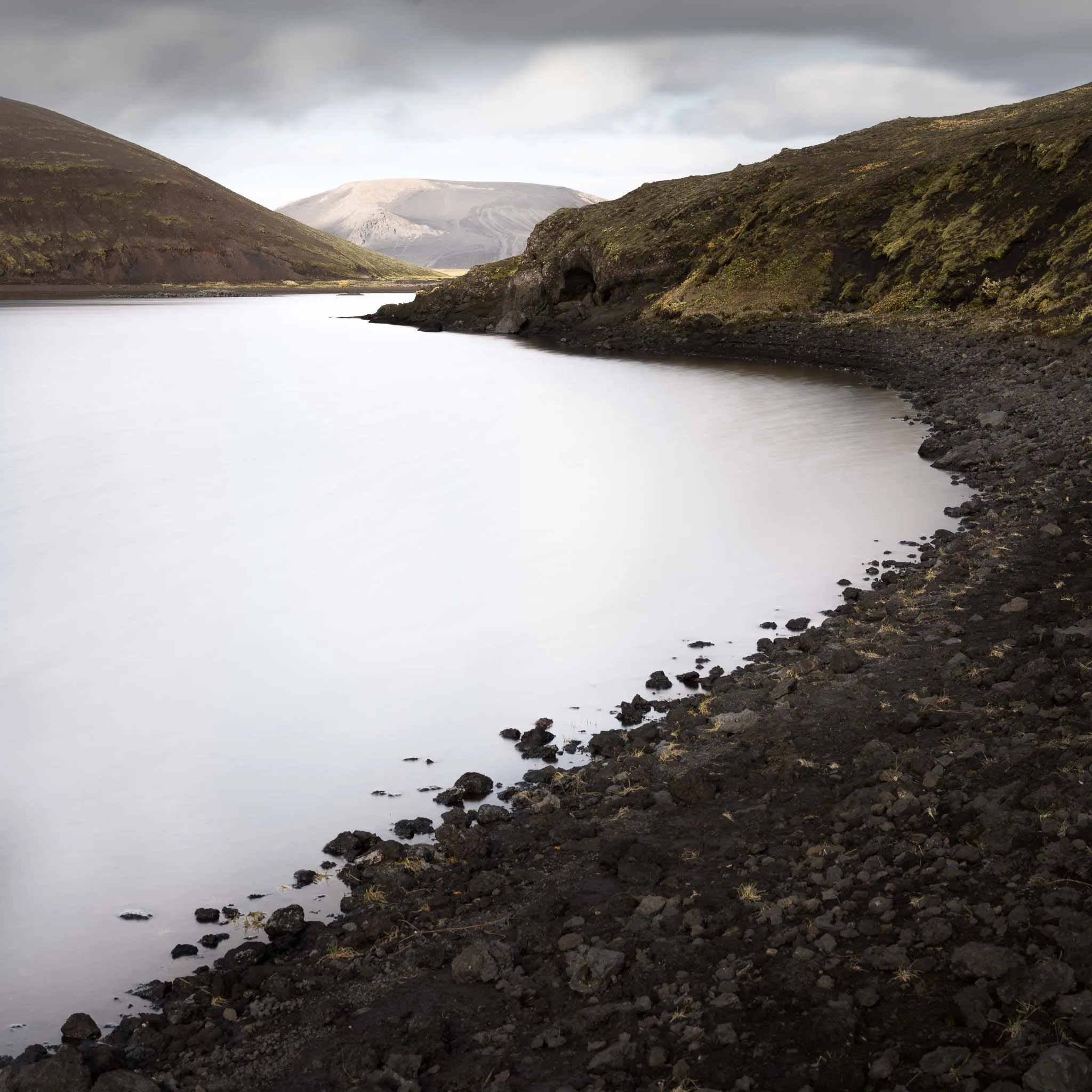A milky, silky lake curving between black volcanic slopes with a pale mountain beyond