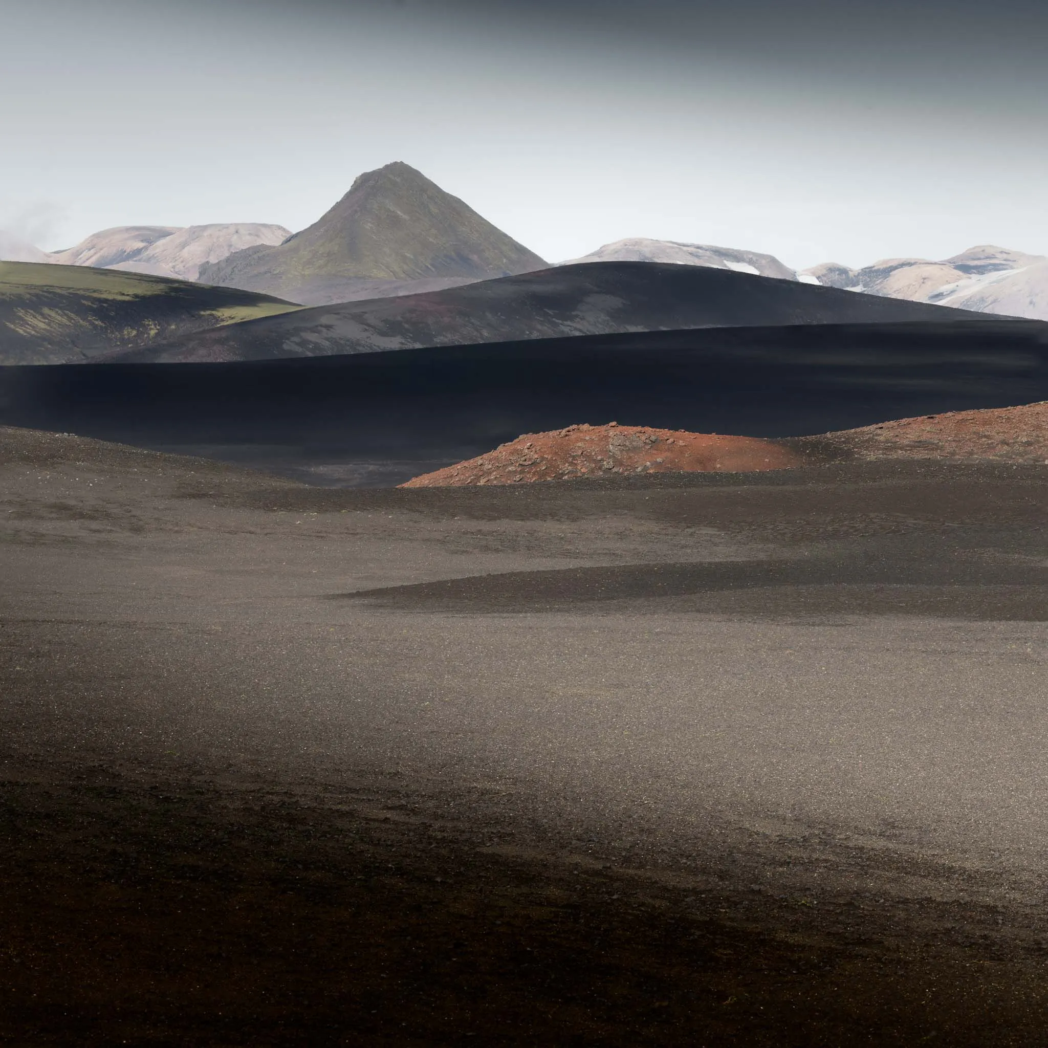 A black volcanic desert with a red cinder mound and green-tinged hills below a peak