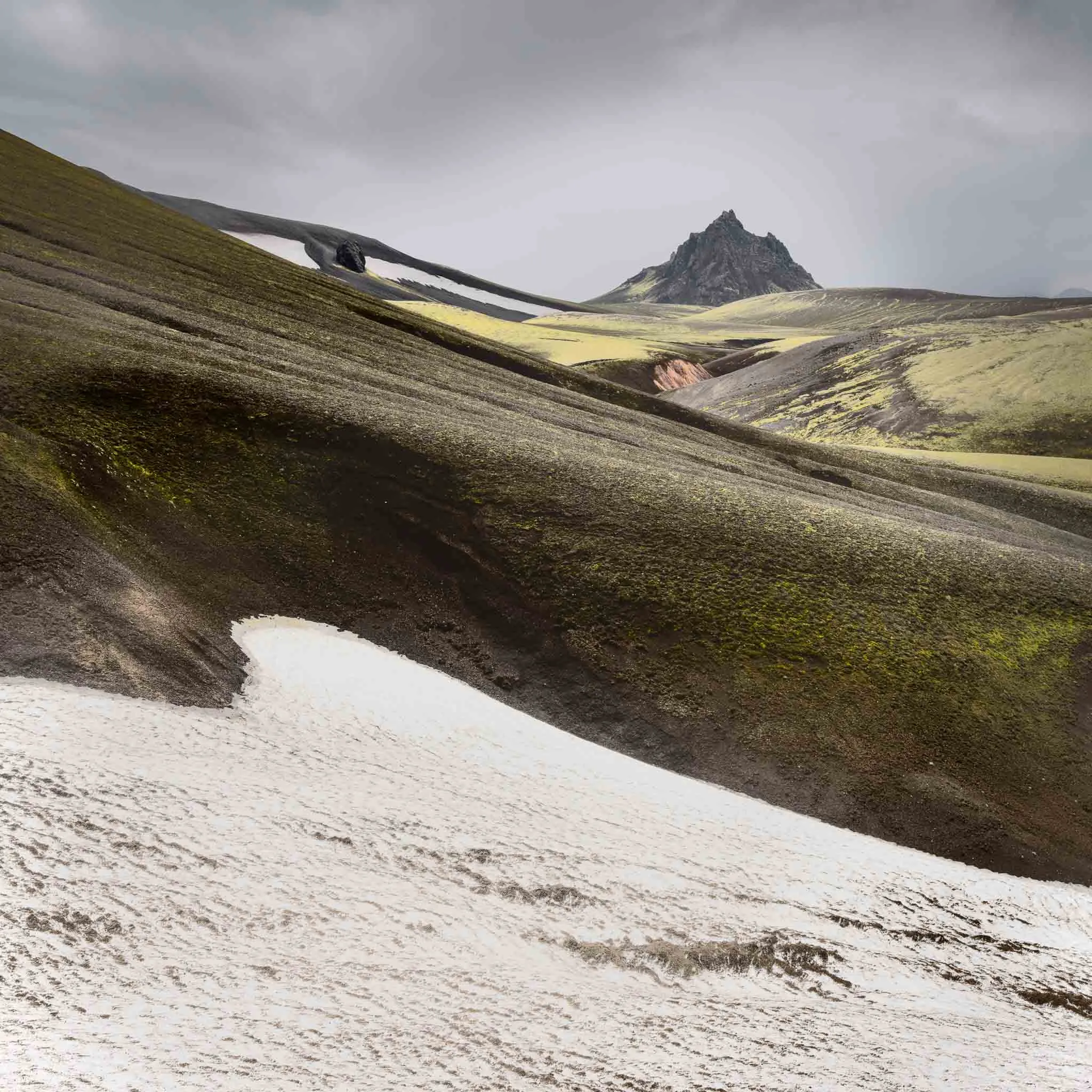 Moss-covered volcanic hills with a snow patch and a jagged dark peak under a grey sky