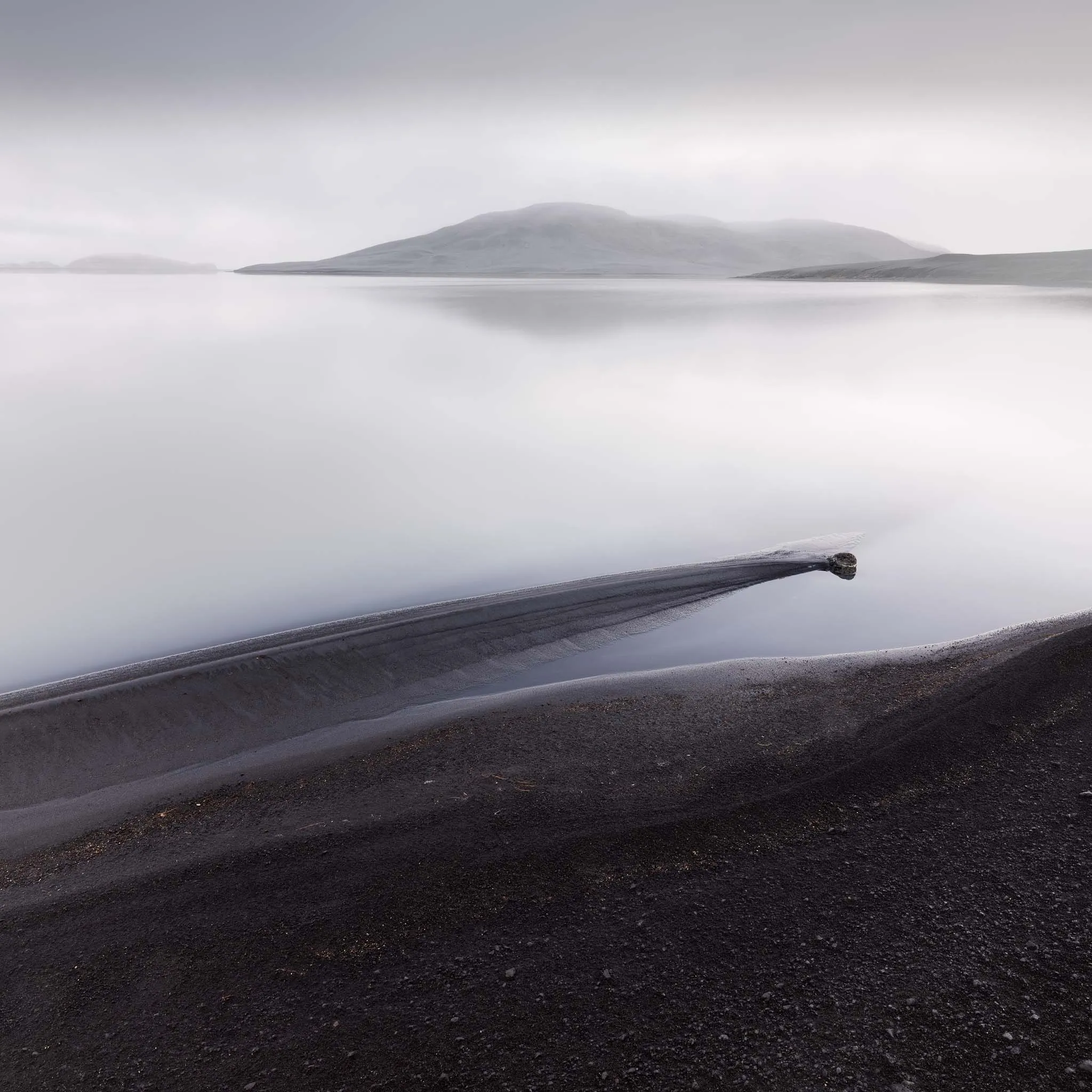 A black sandbar reaching into a still, misty lake below soft hills