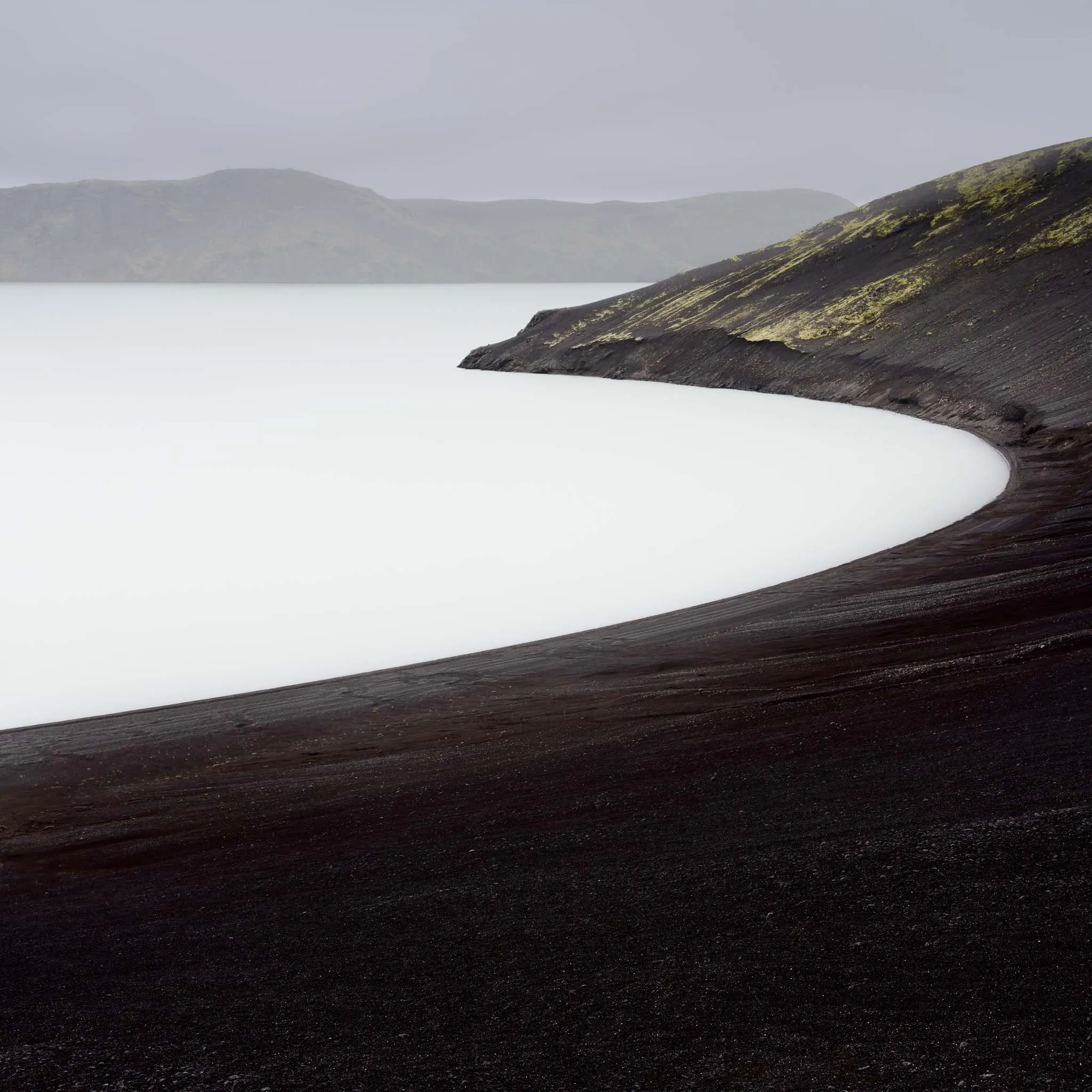 A pale, milky lake ringed by black volcanic ash slopes with misty mountains beyond