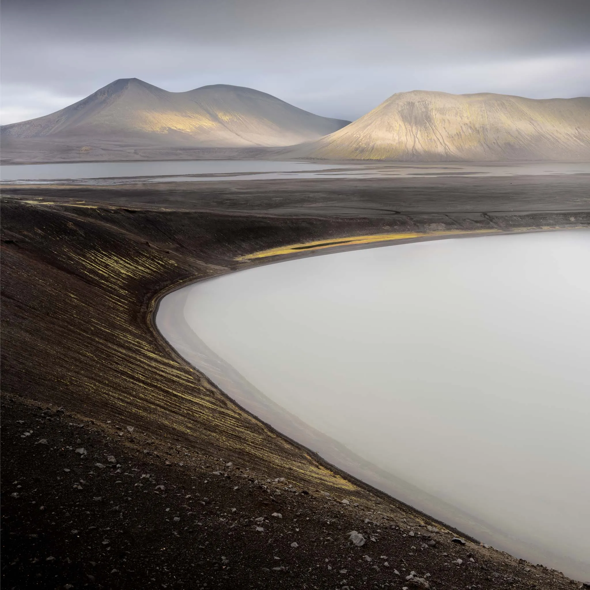 A curving pale lake edged by black ash, cone-shaped mountains catching light behind