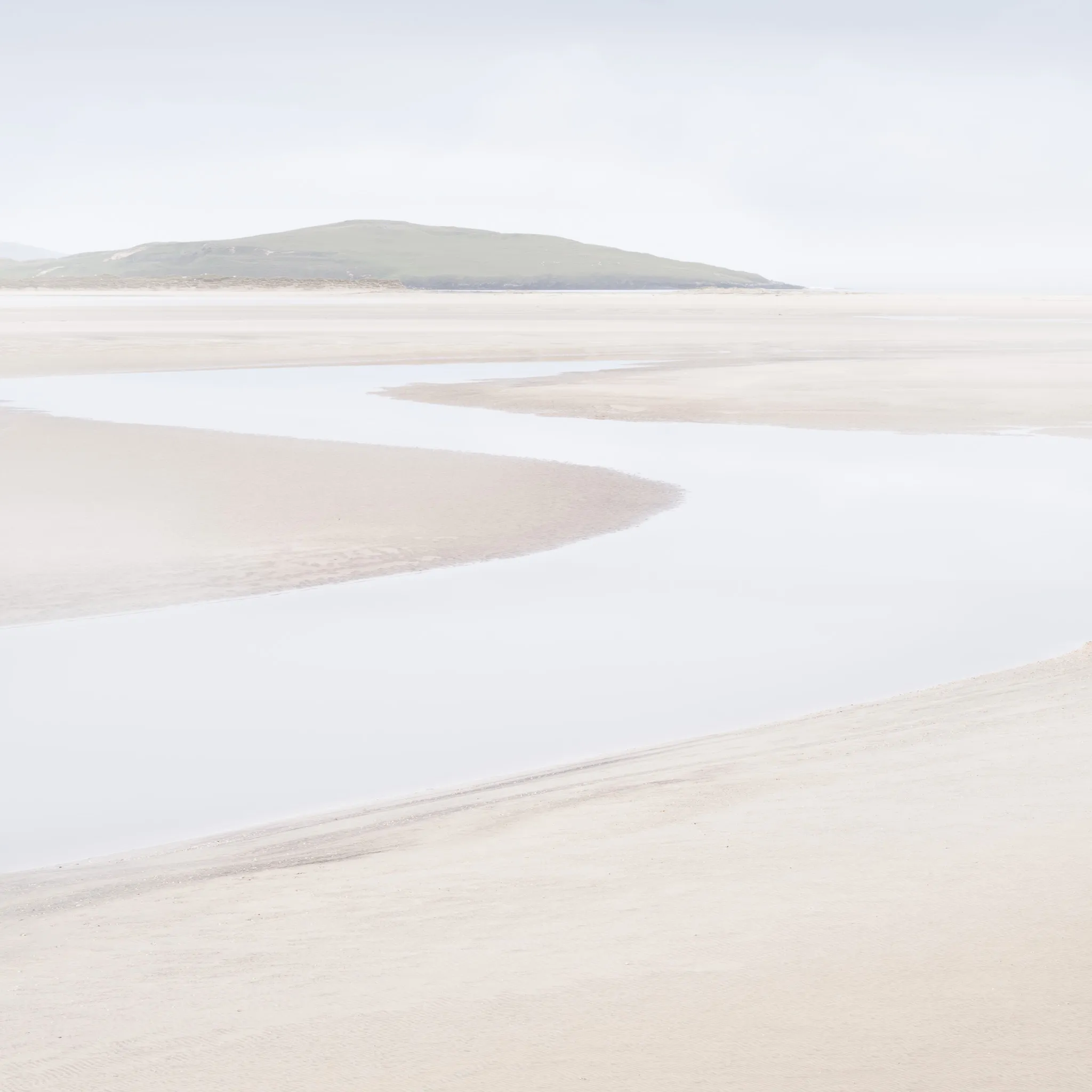 A winding tidal channel across a pale, empty beach with a green hill beyond