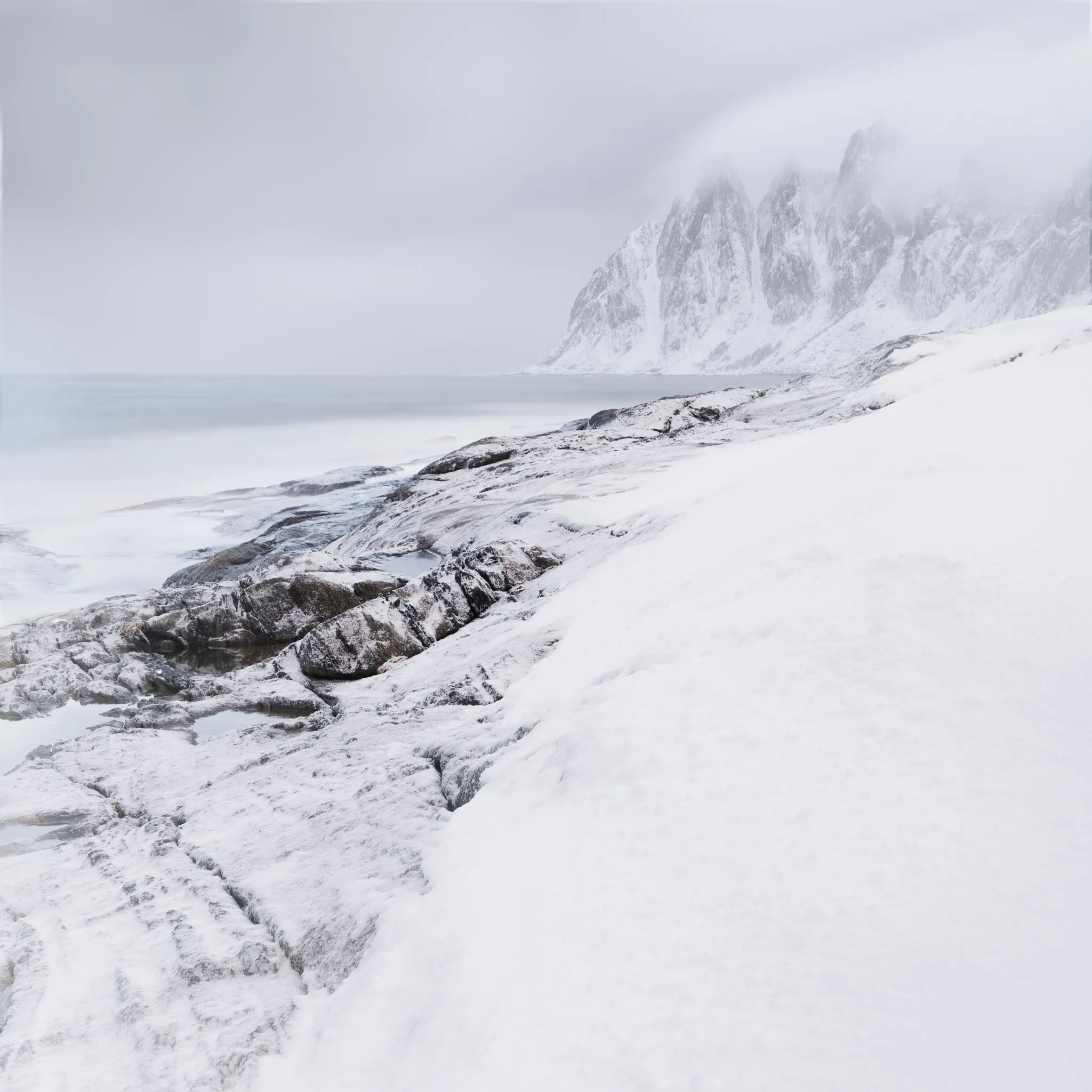 Snow-covered coastal rocks below the jagged Okshornan peaks fading into mist above a calm sea