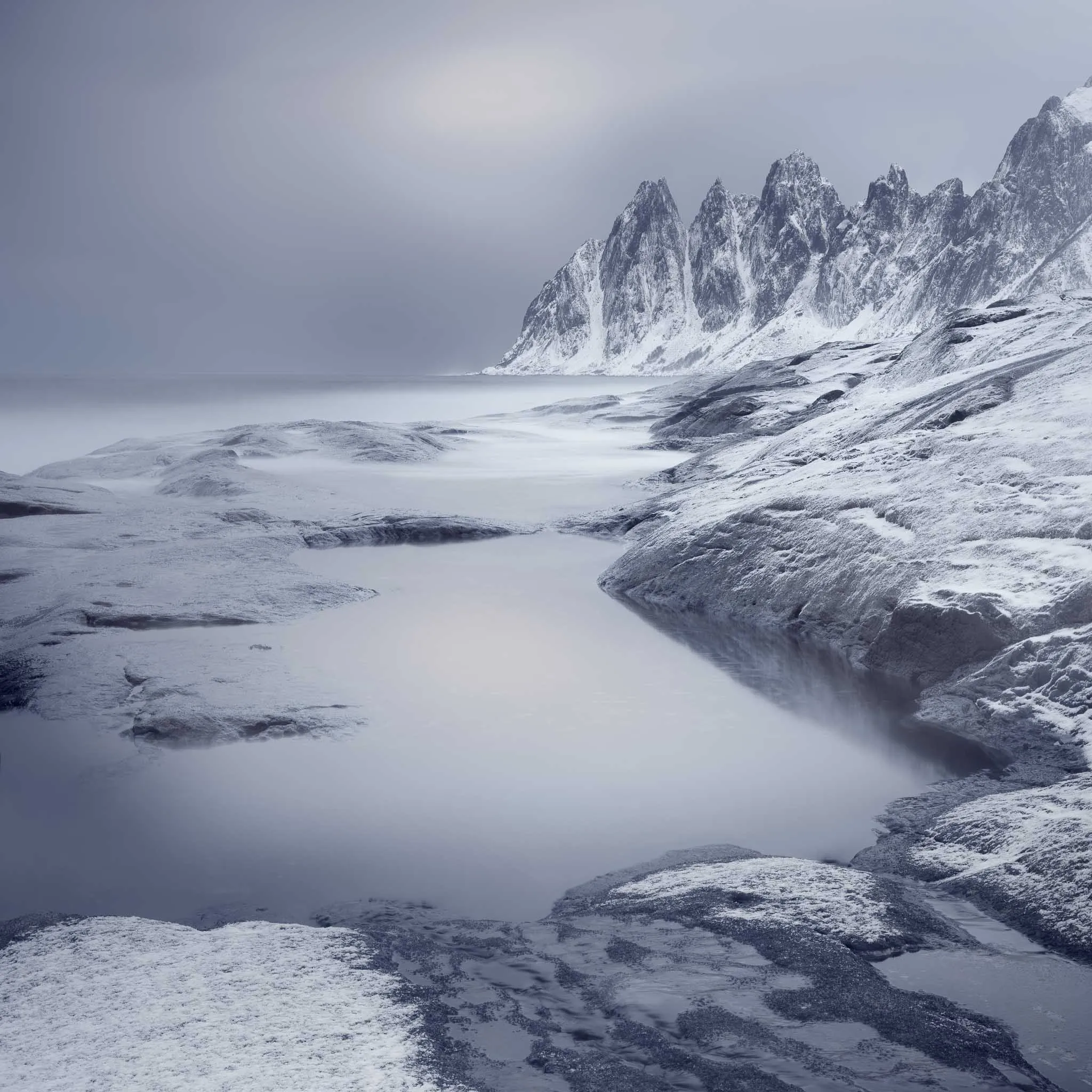 Sharp snow-clad peaks rising beyond a smooth, misty bay and a quiet tidal pool