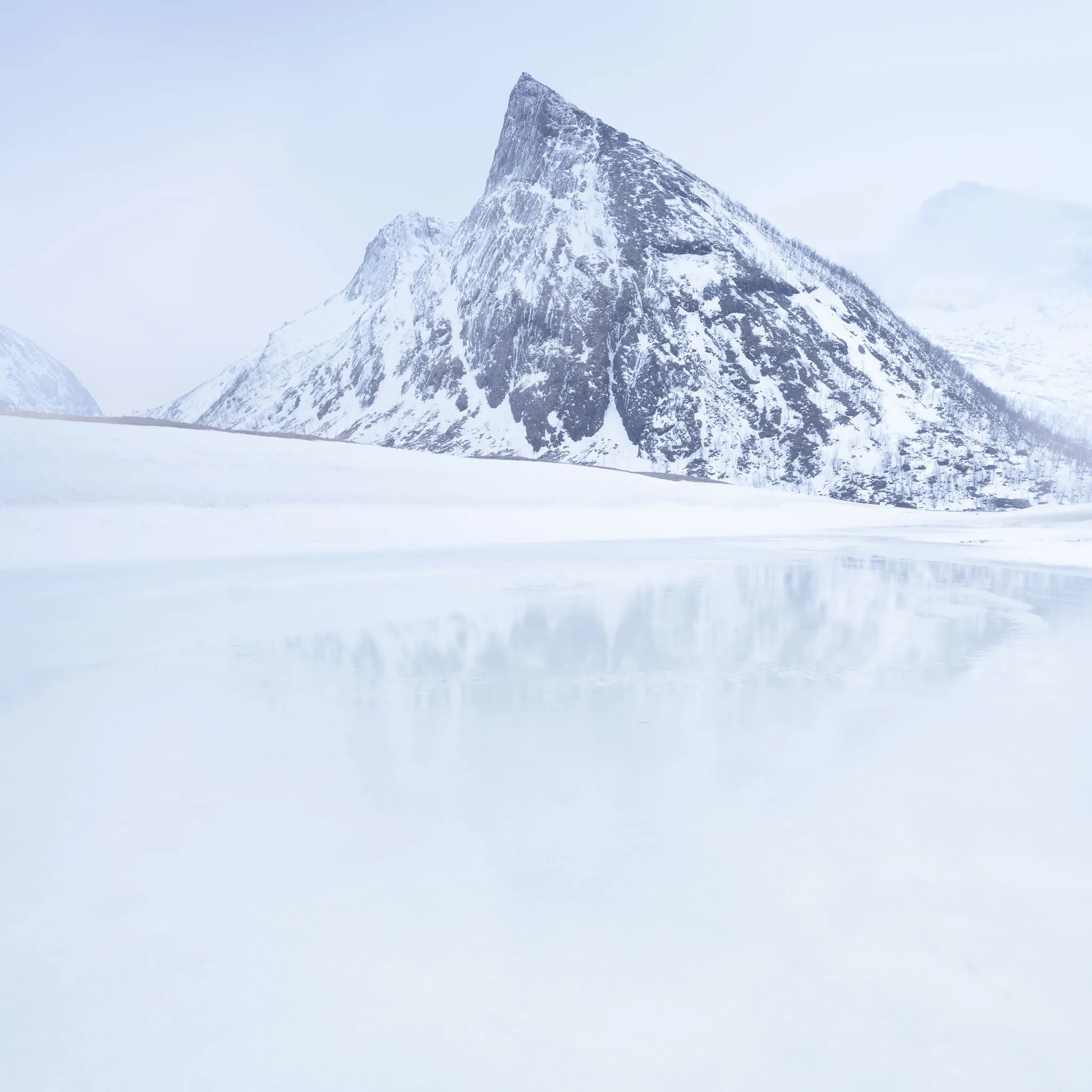 A dark pyramidal peak above a frozen lake in soft high-key light