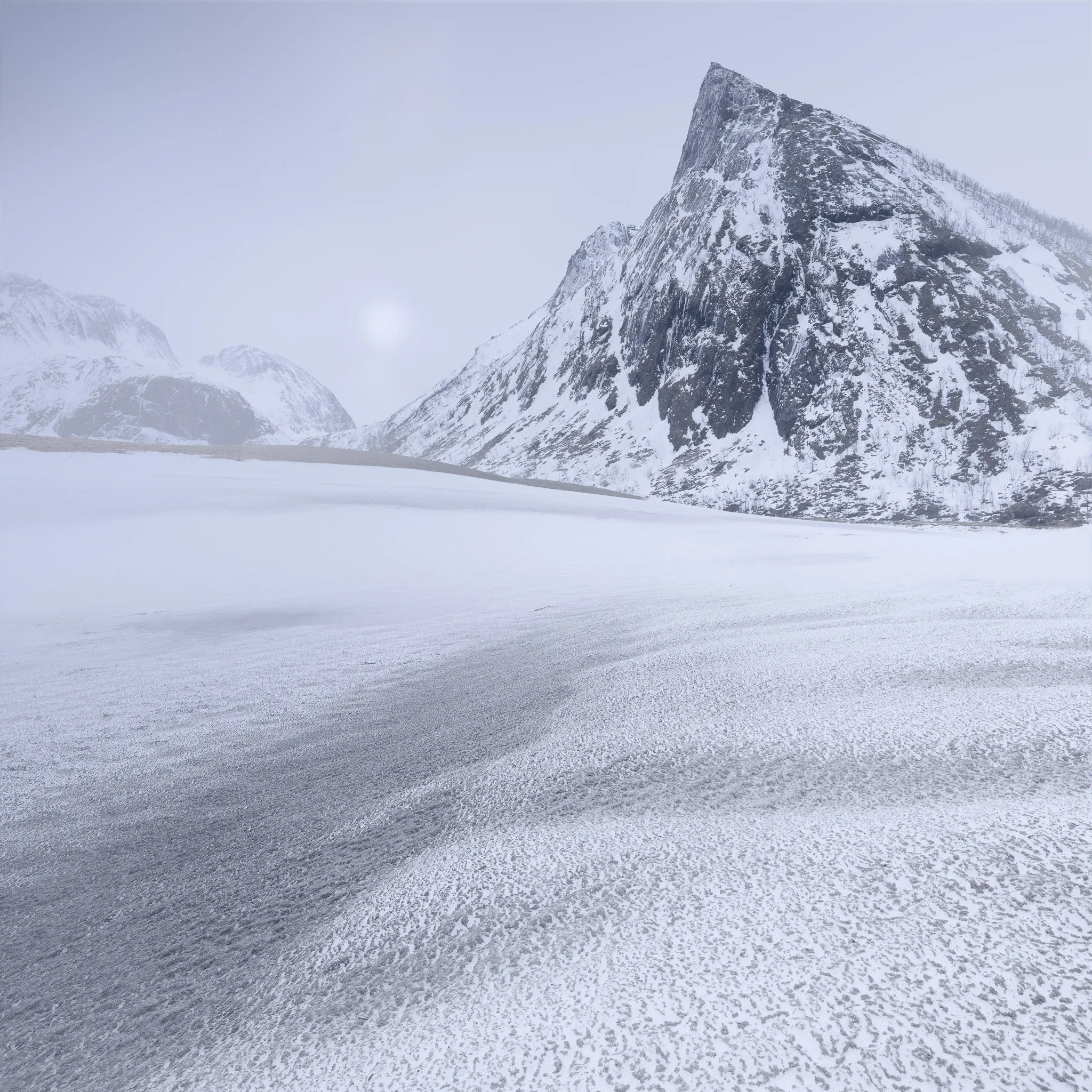 A pale sun behind cloud over a pyramidal mountain and a wind-textured frozen lake