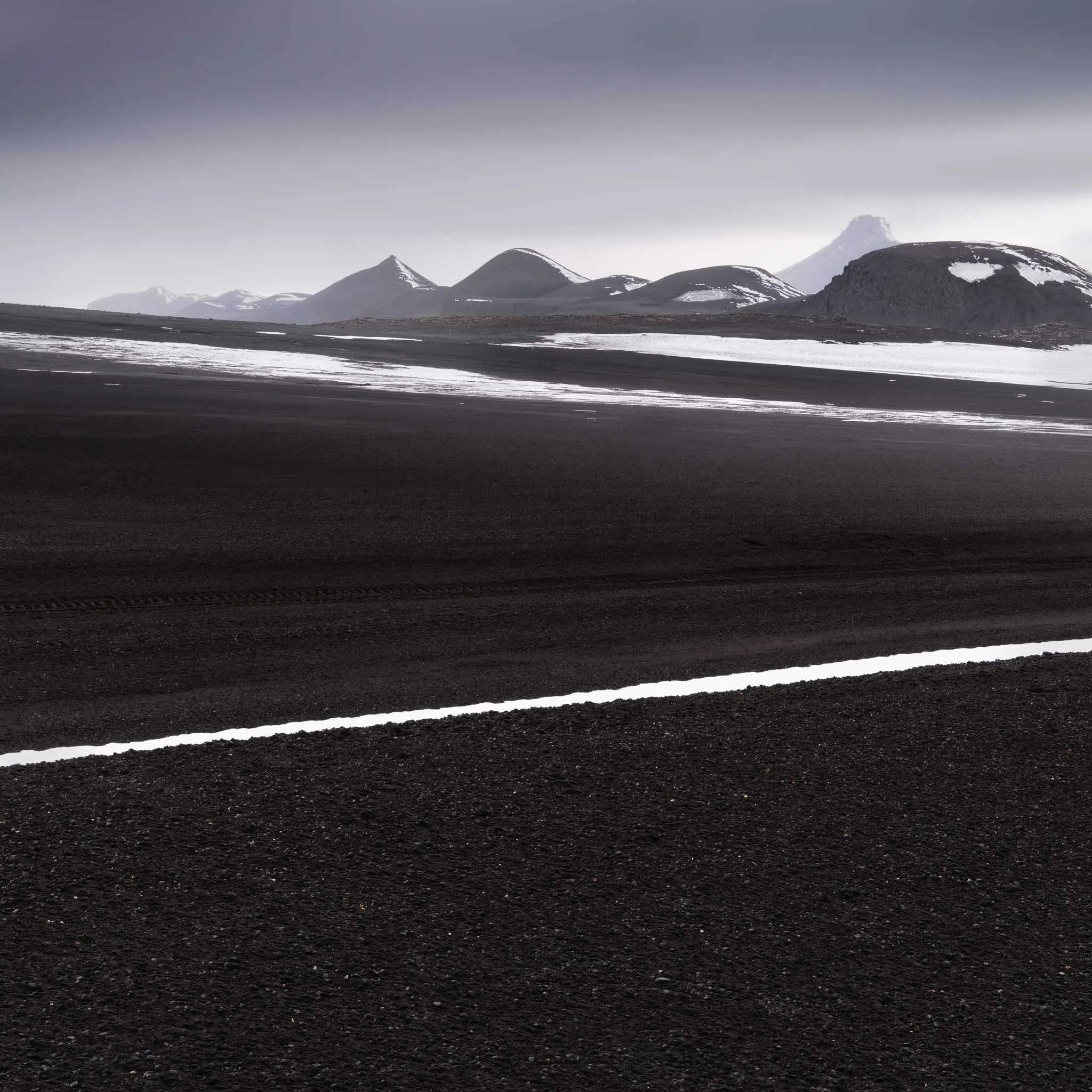 A vast black volcanic plain streaked with thin snow lines, snow-dusted mountains and a jagged peak beyond