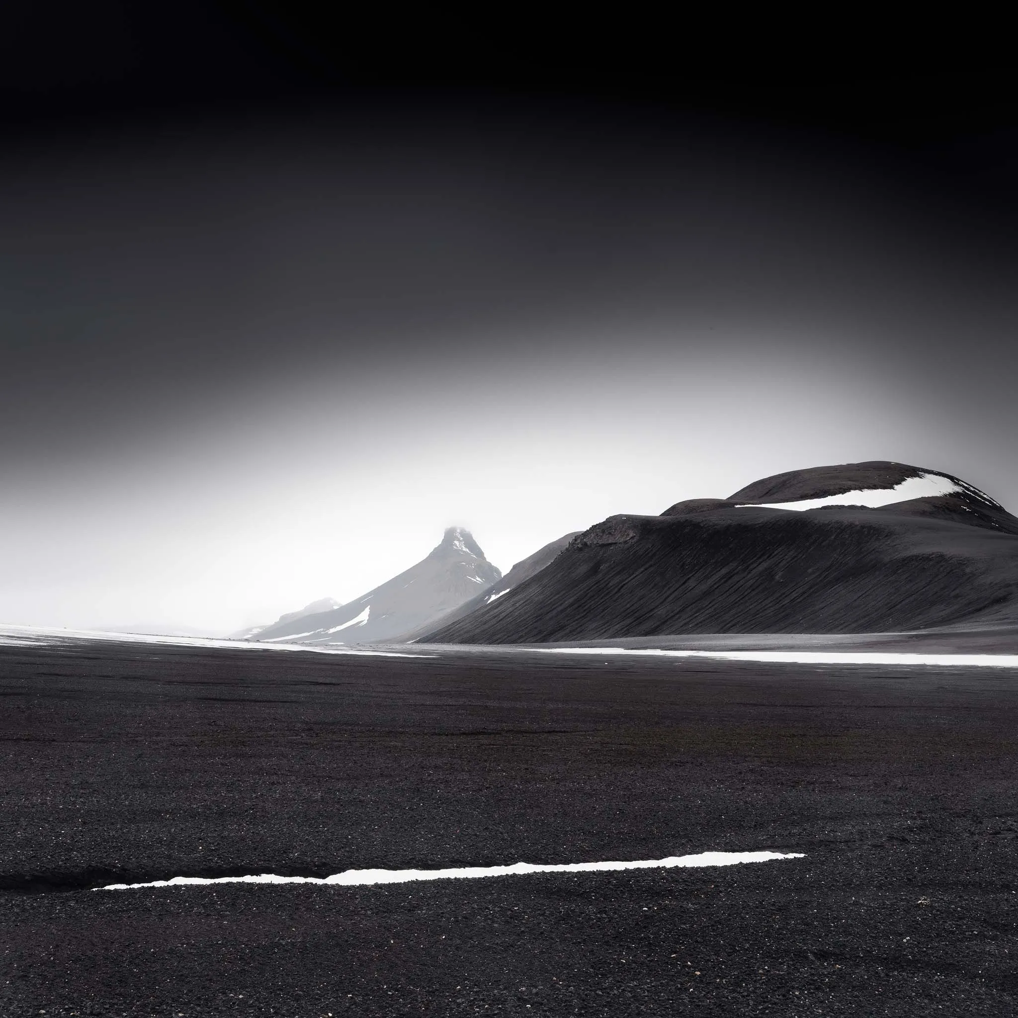A black ash plain with a curving snow line below a horn-shaped peak and dark hill