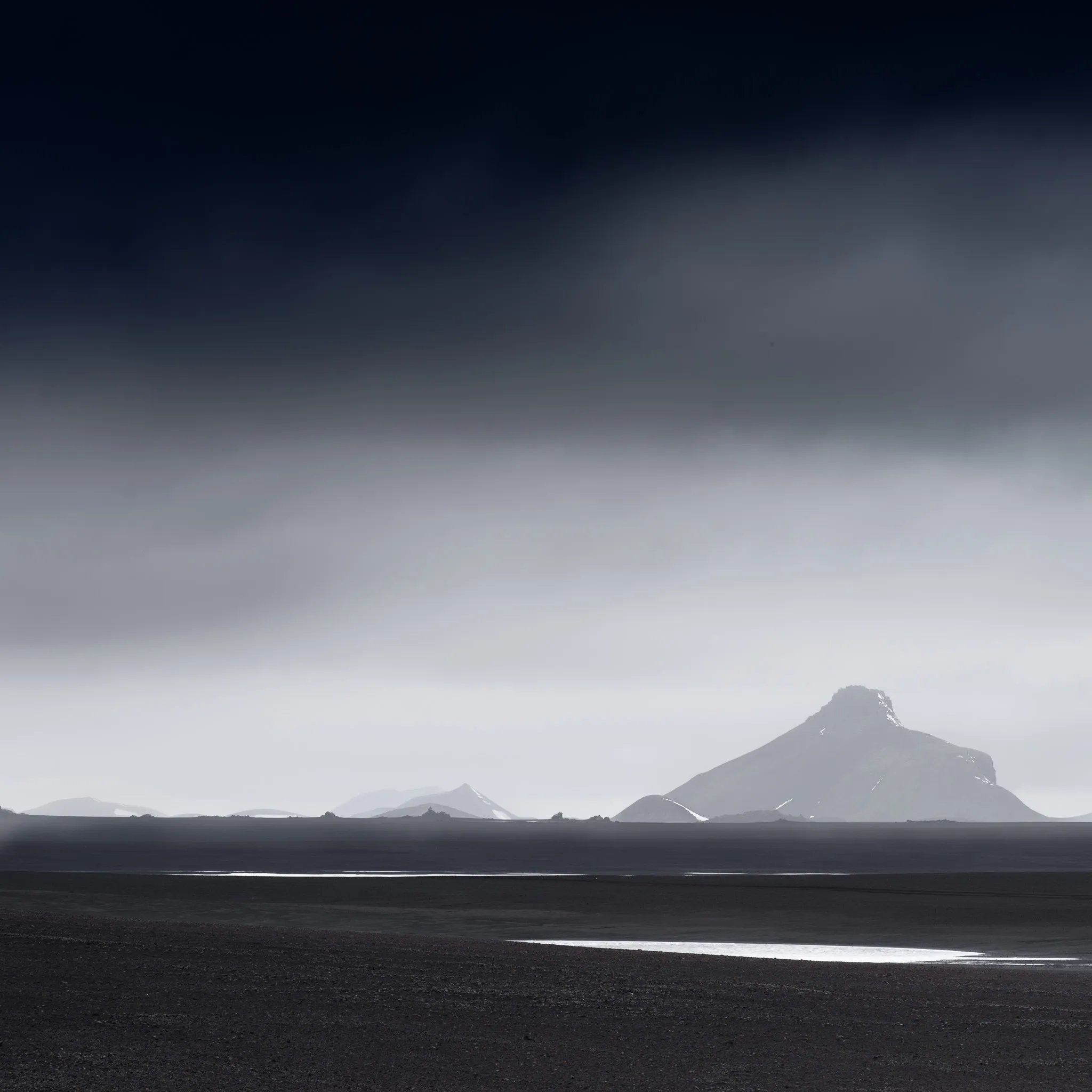 A solitary horn-shaped peak across a black desert under a heavy dark sky, a bright pool of meltwater