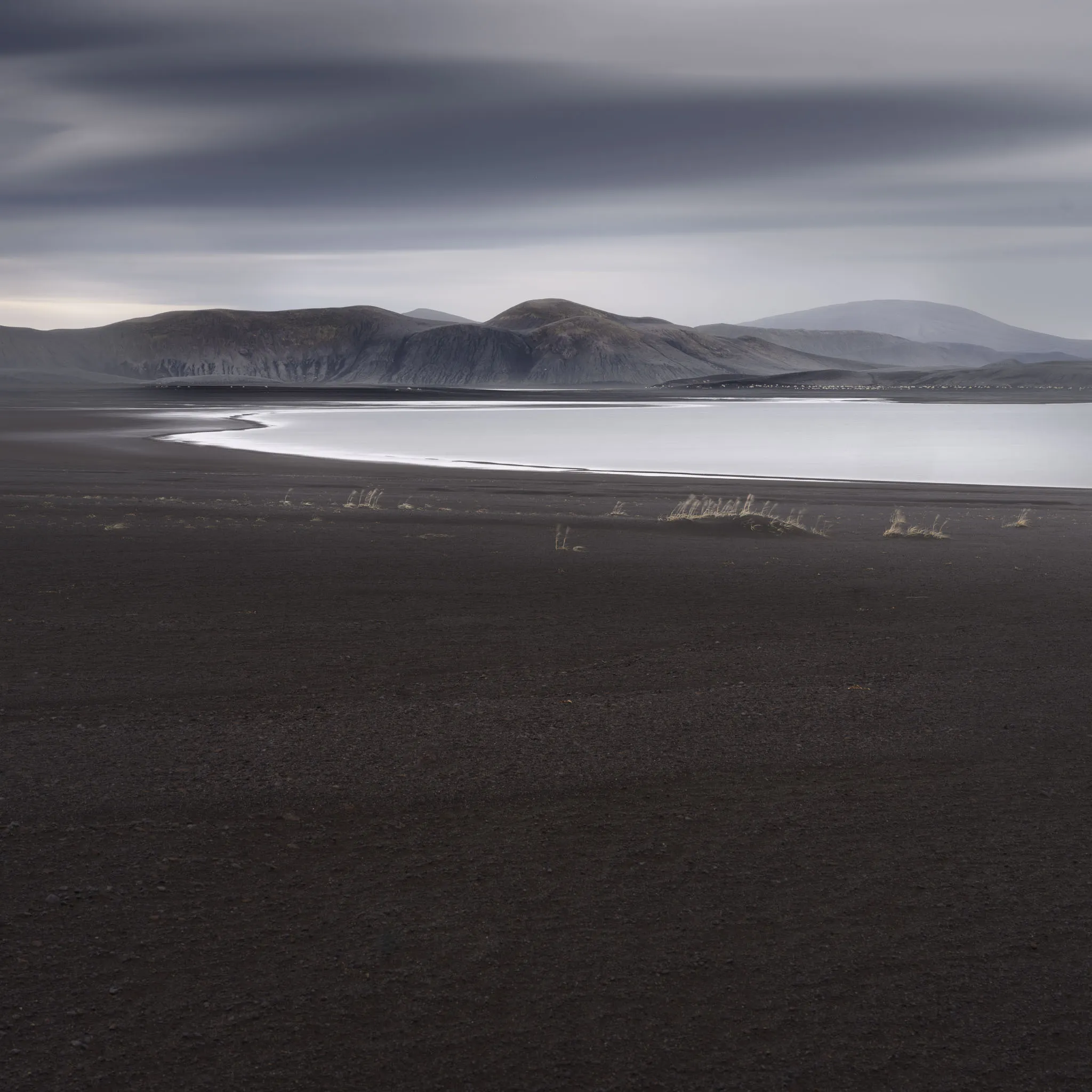 A black-sand plain with sparse tufts of grass, a pale lake and dark mossy mountains beyond