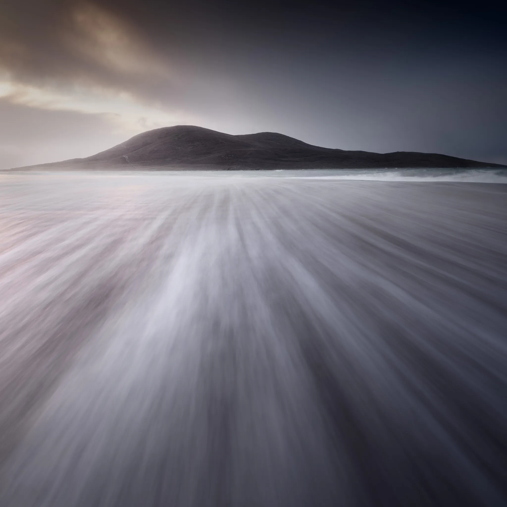 Wind-blown sand streaking across a beach below a dark hill under a moody sky