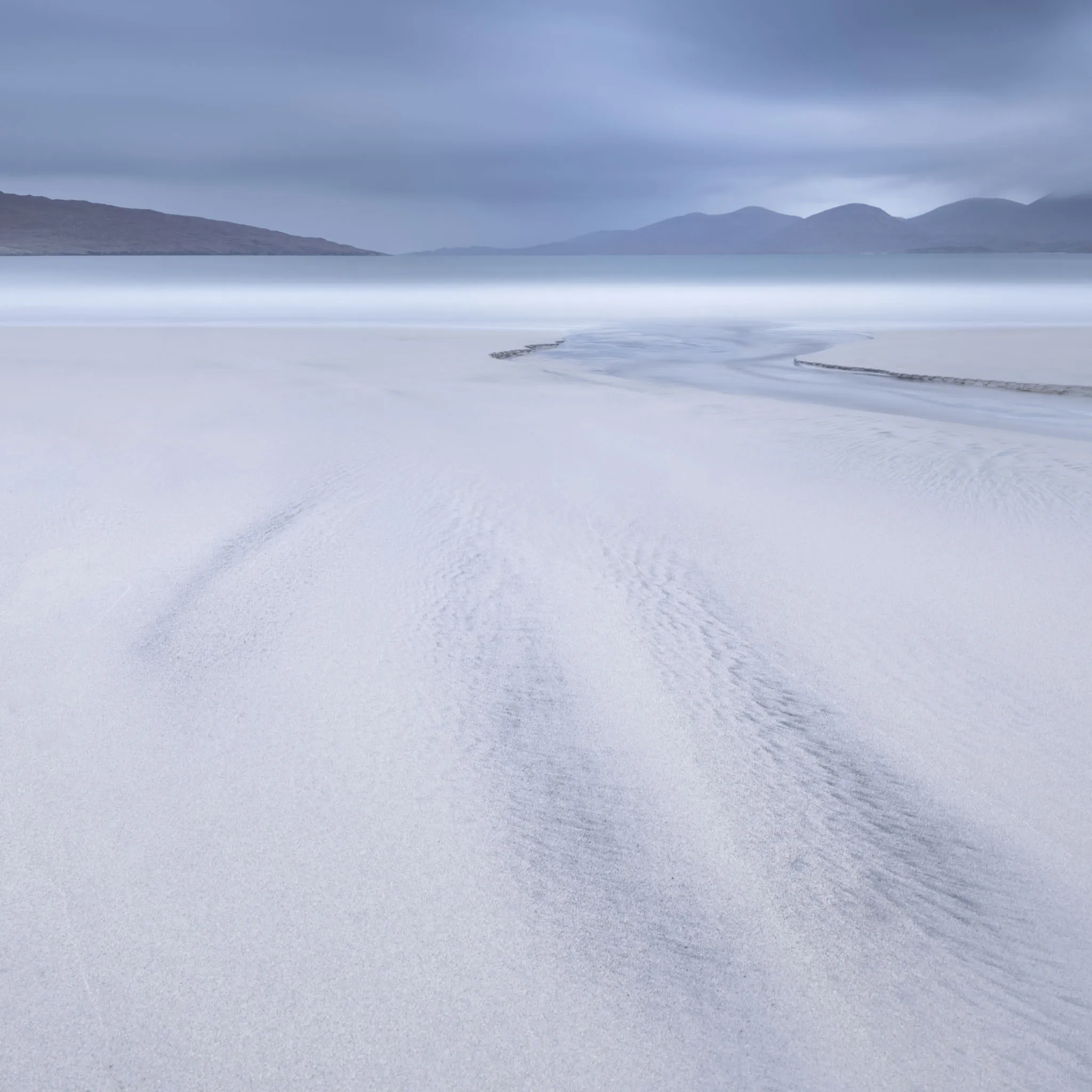 A cool blue beach with a curving tidal channel and hazy mountains behind