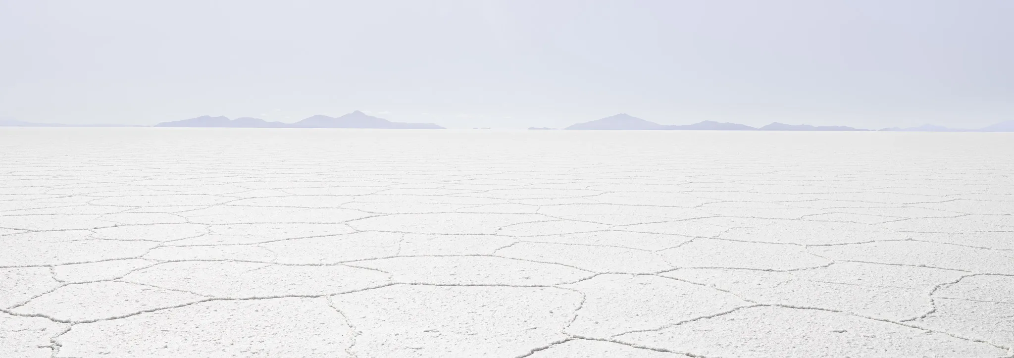 The cracked hexagon crust of a salt flat stretching to distant blue mountains under a pale lilac sky