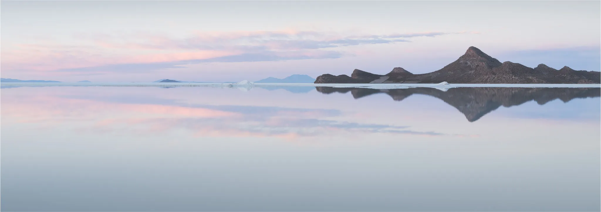 Dark rocky islands mirrored in a flooded salt flat at dusk, soft pink and lilac clouds above
