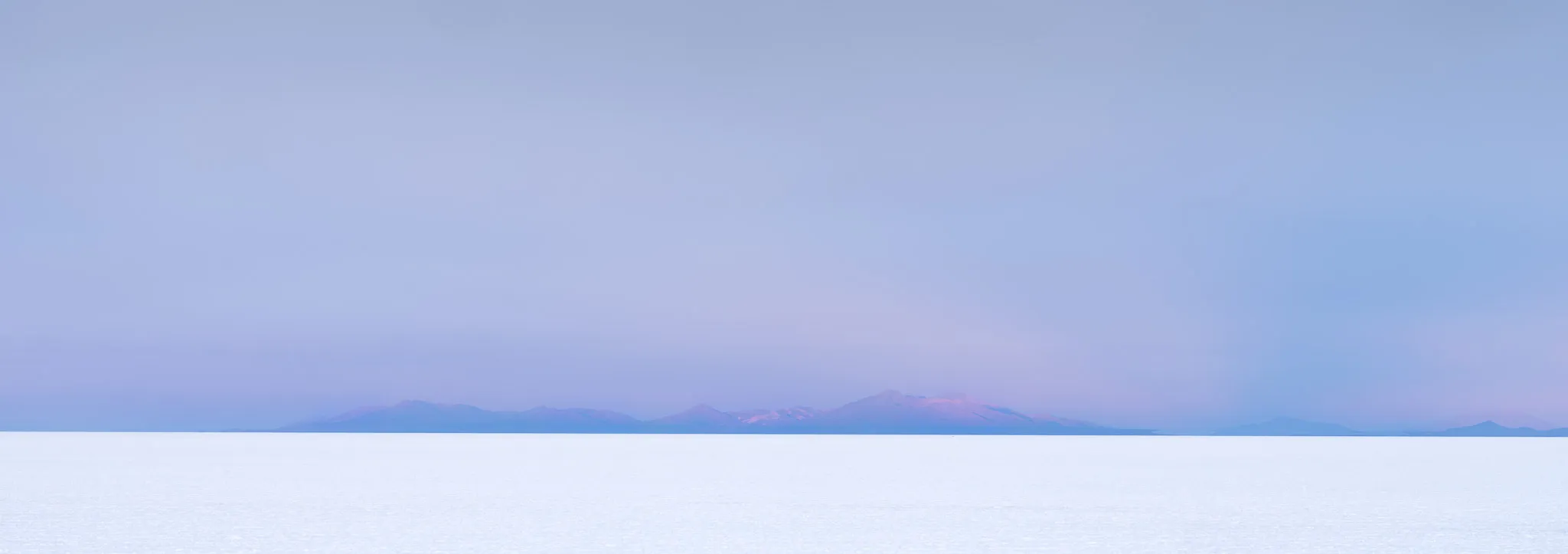 A faint line of distant mountains over a pale, flooded salt flat beneath a lavender twilight sky