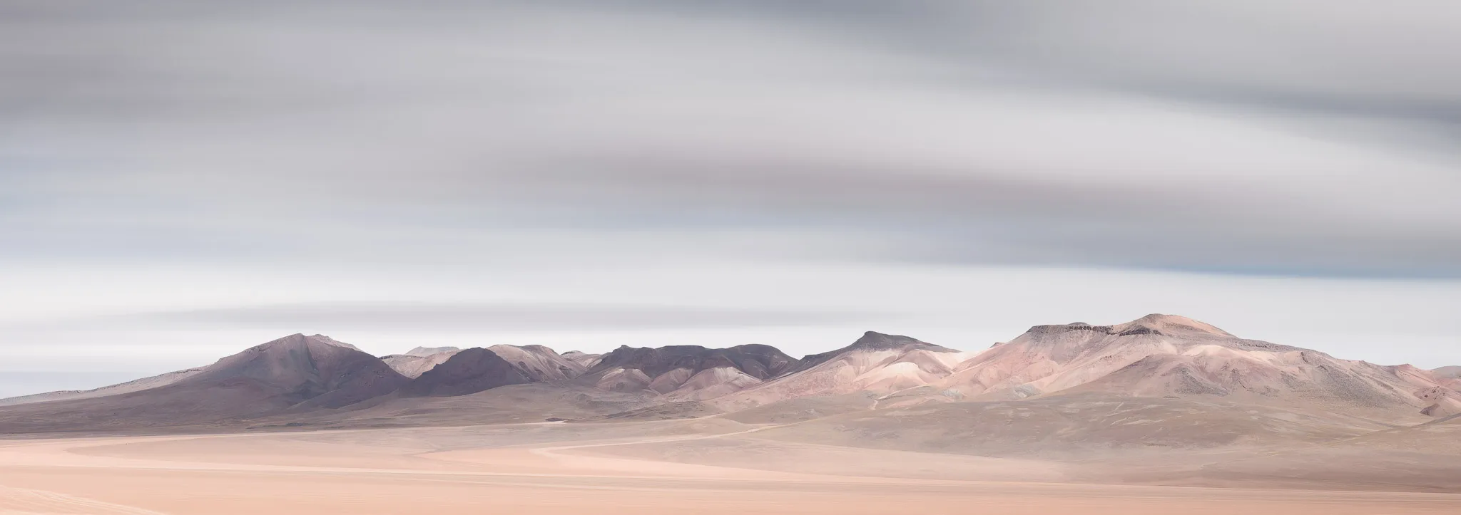 A pale high-desert plain sweeping toward layered pastel mountains under a soft, streaked sky