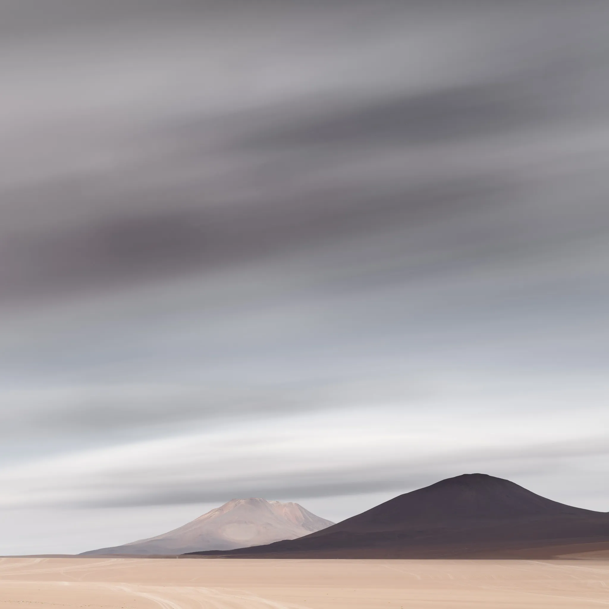 Two volcanic cones, one pale and one dark, rising from a wide desert plain beneath streaked, slow-moving clouds