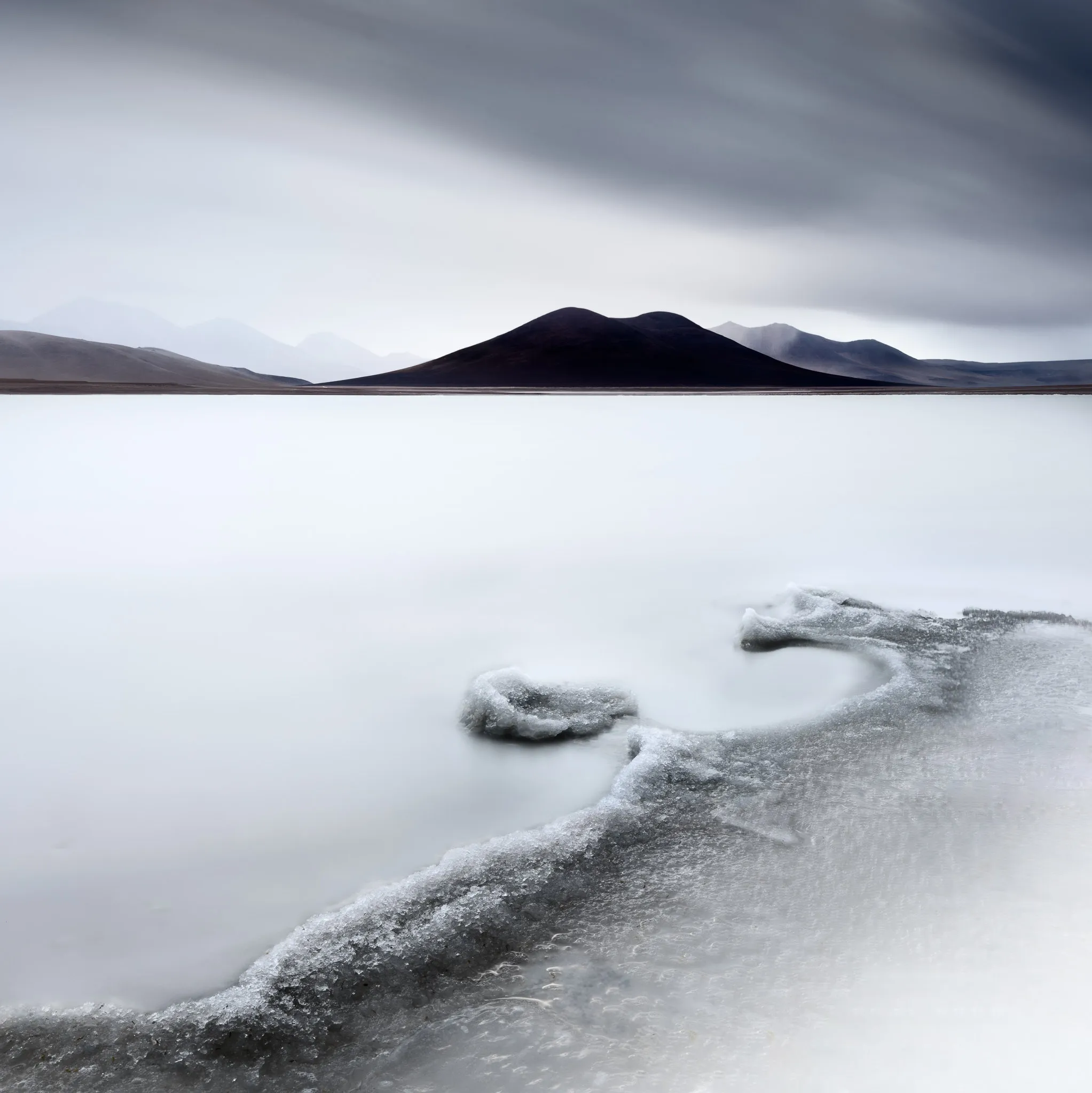 A salt-crusted lagoon with milky, still water below a dark solitary mountain and layered peaks