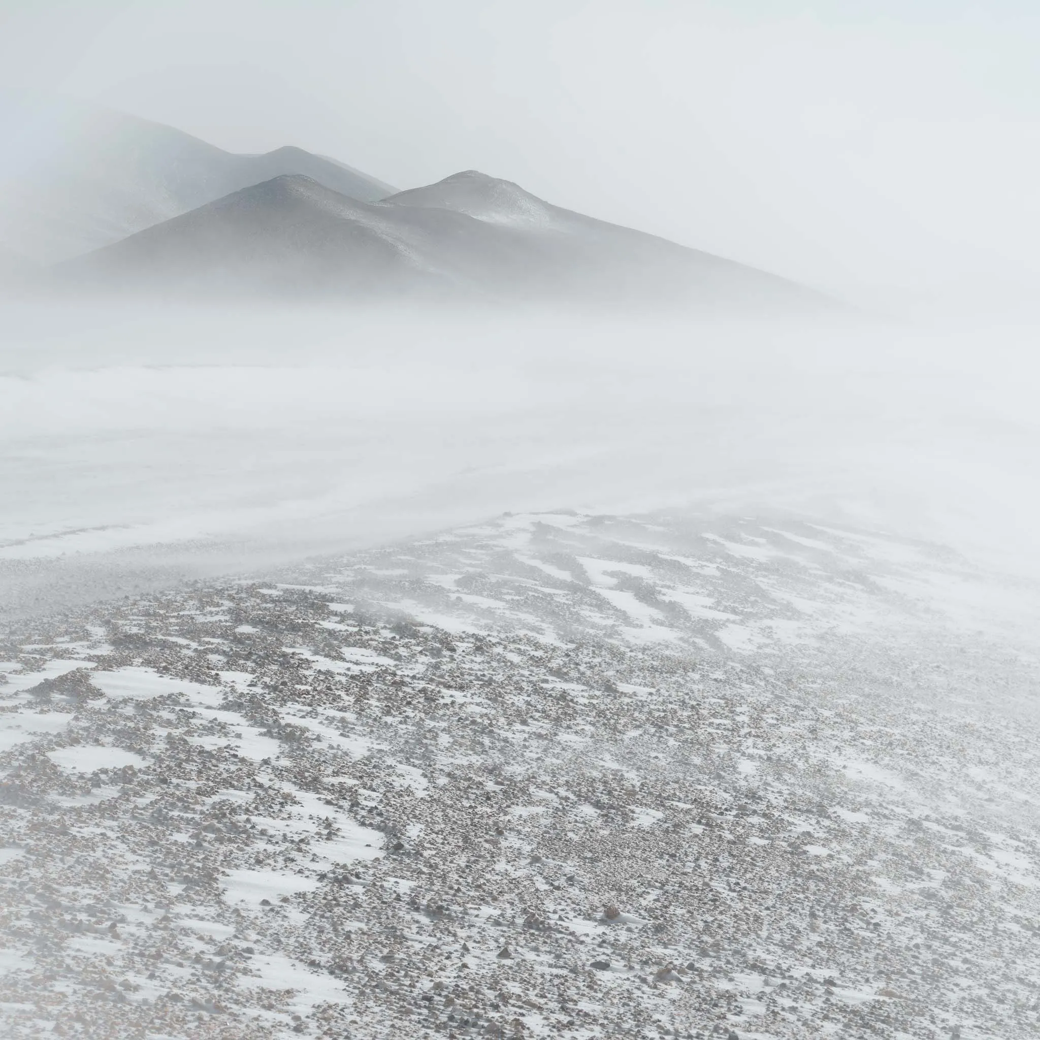 Snow-veiled mountains looming through blowing mist above a rocky, snow-streaked foreground