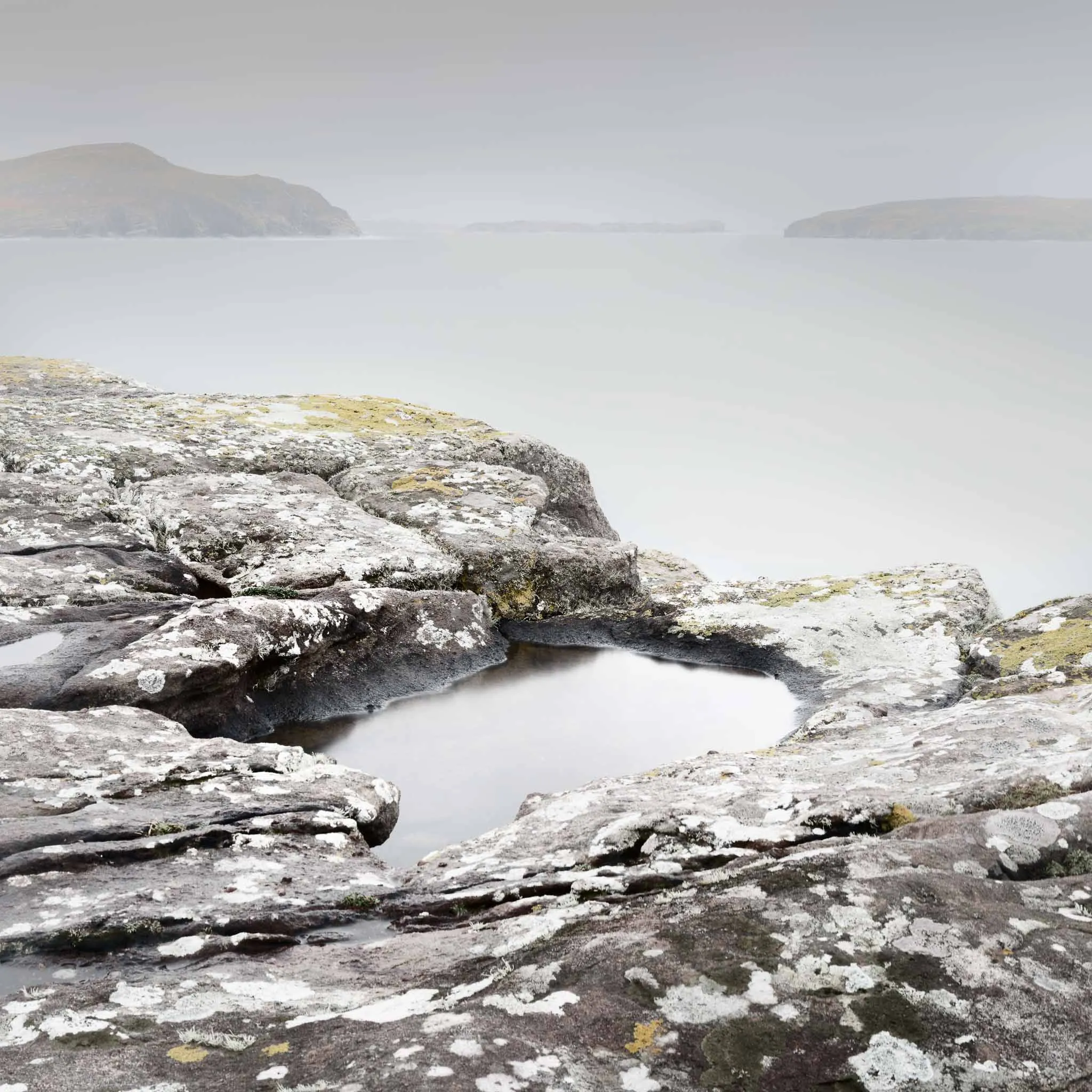 Lichen-mottled coastal rocks around a still pool, misty headlands receding across a pale sea