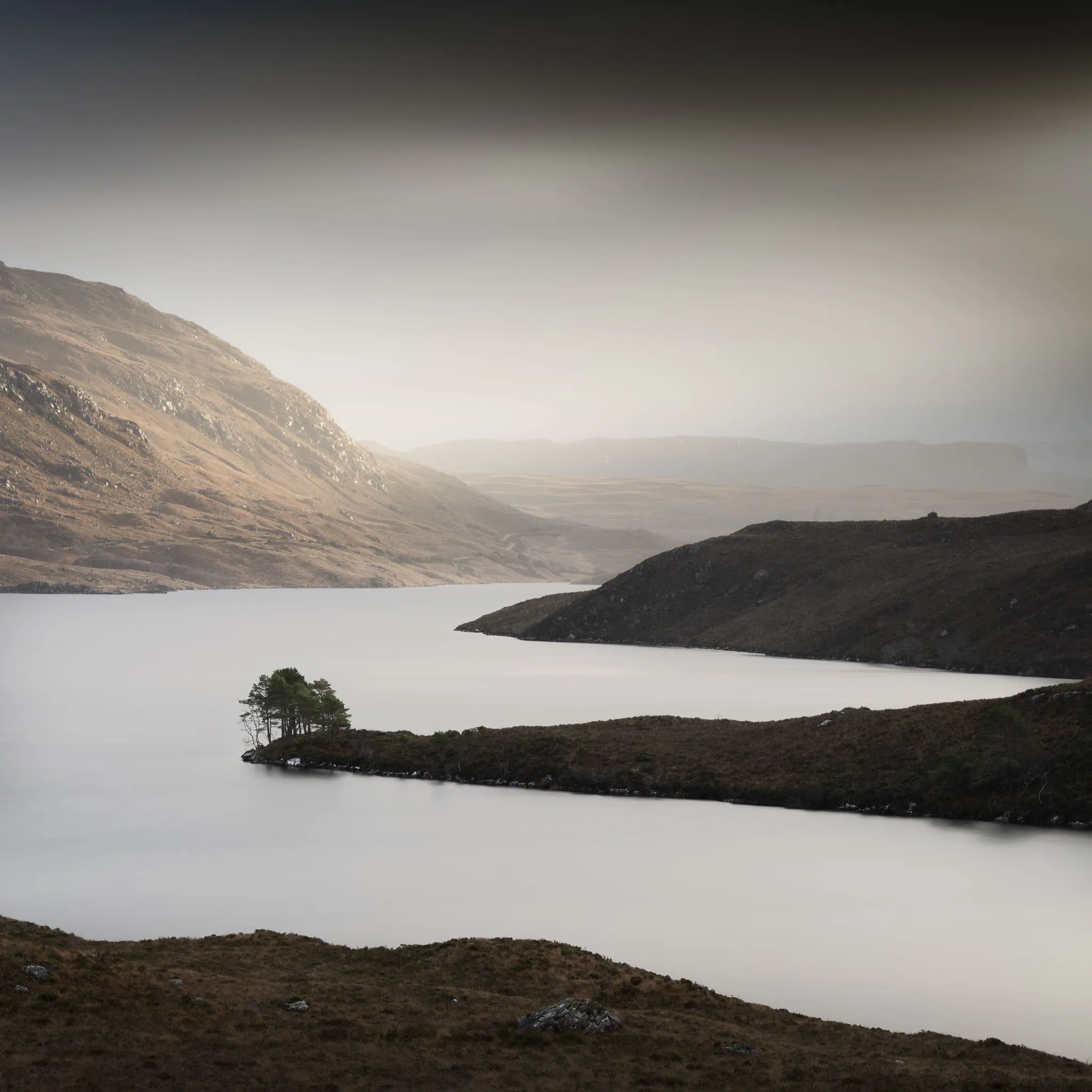Warm light over Loch Assynt, a tree-topped promontory reaching into calm water below tawny highland hills