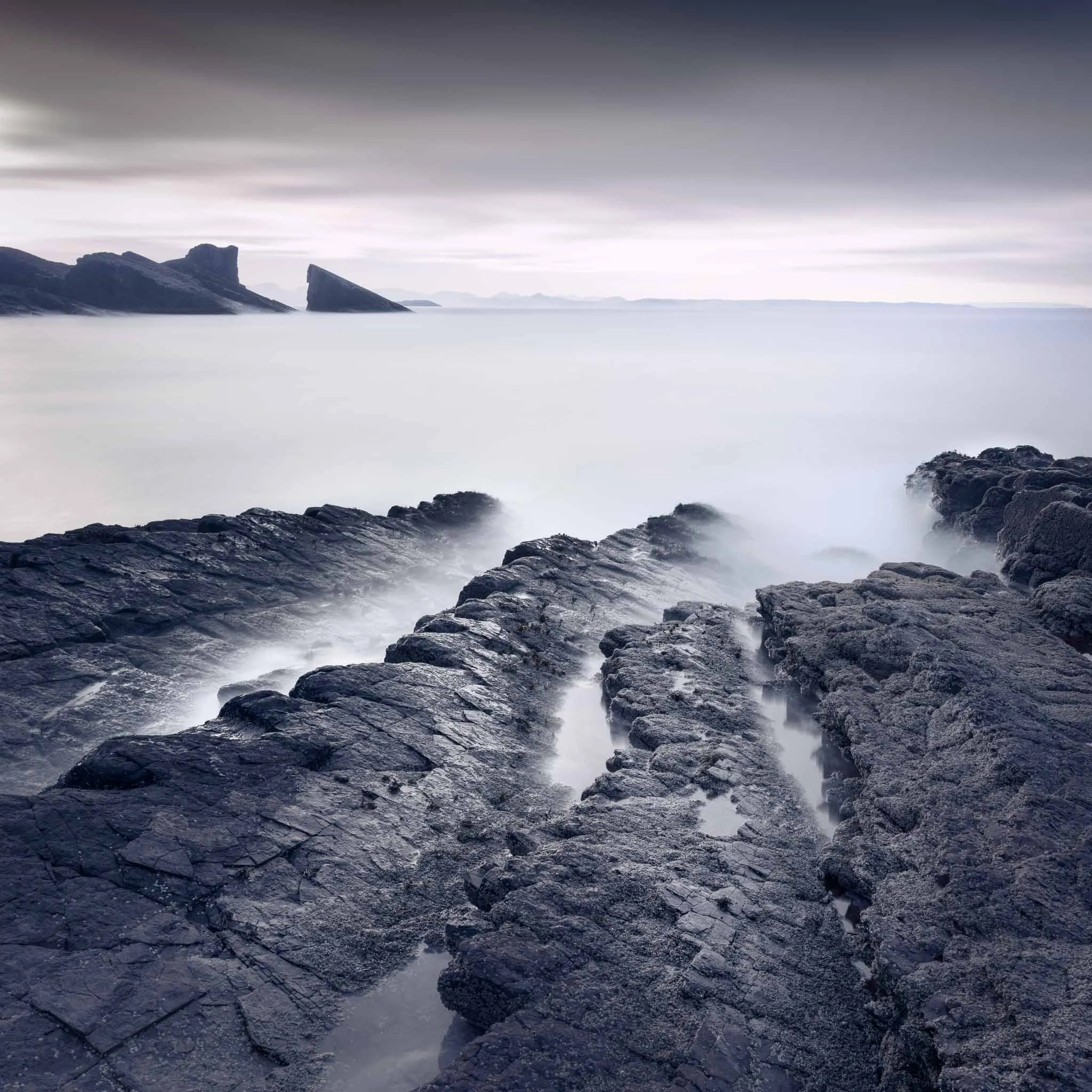 Dark, weathered coastal rock ledges running out toward jagged sea stacks under a mauve twilight sky