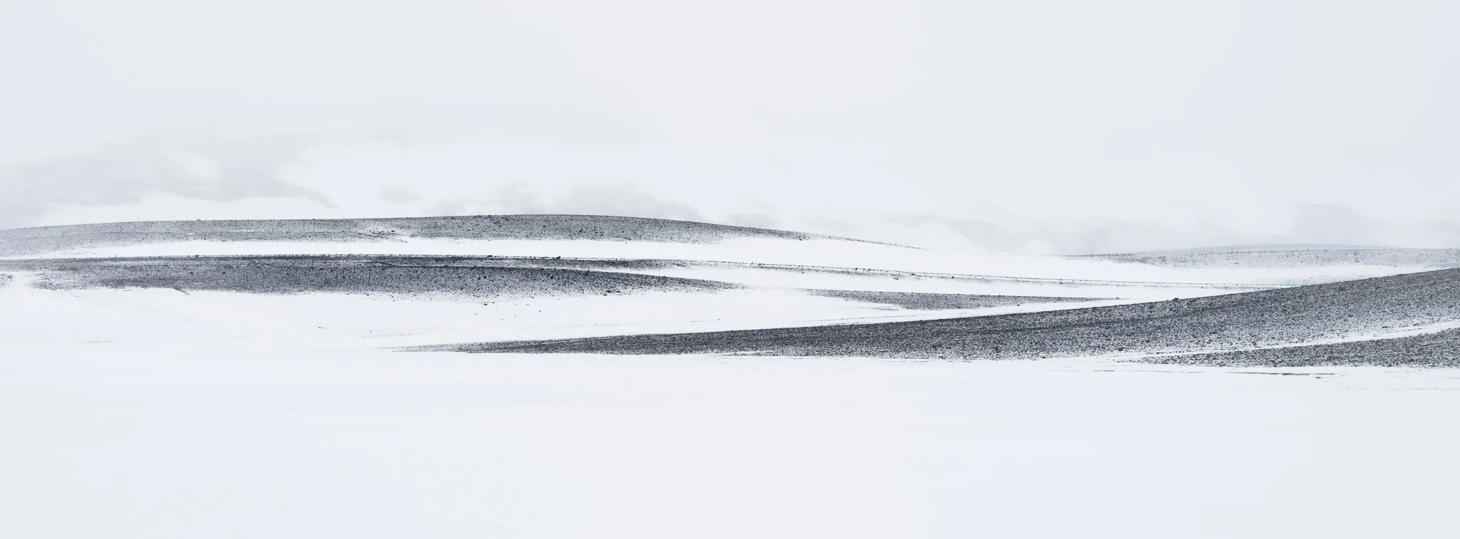 Dark ground curving through pristine snow in a near-white Icelandic highland, nearly monochrome