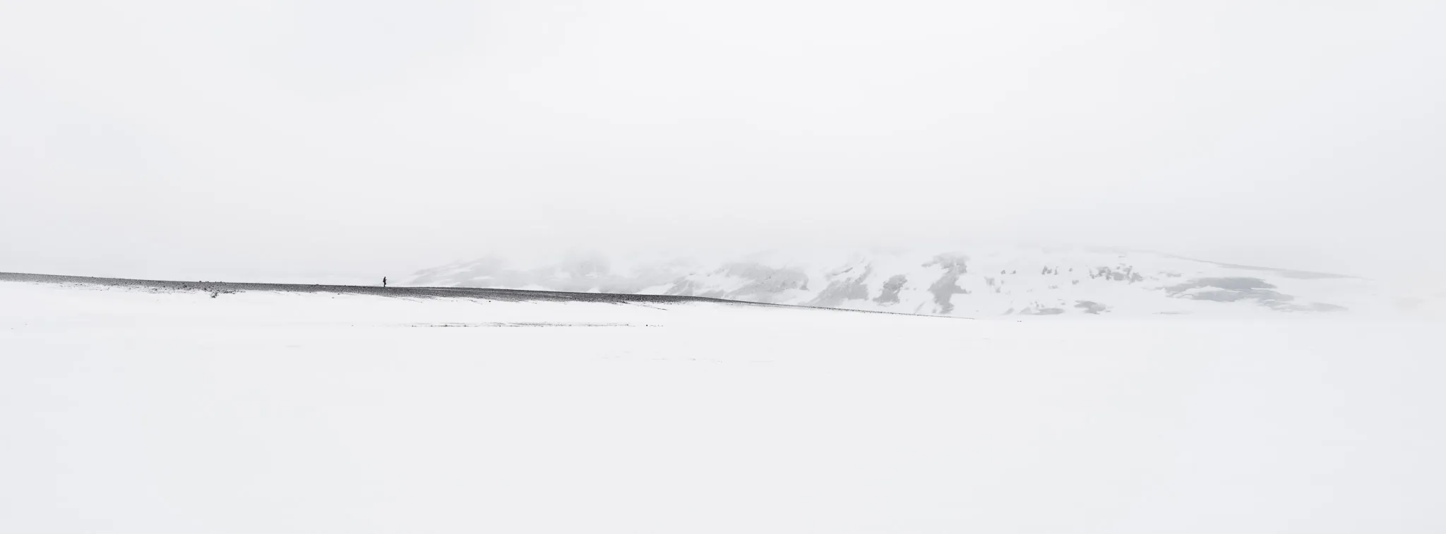 A tiny lone figure on a vast snowy plain below a faint, mist-veiled snow mountain