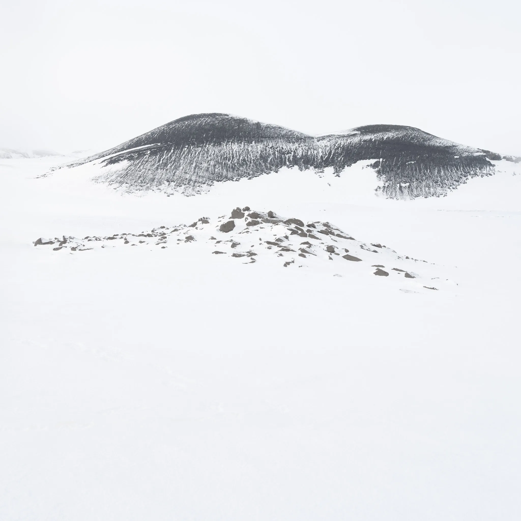 A black, twin-humped hill streaked with snow rising beyond a low mound of dark rubble on a vast white snowfield