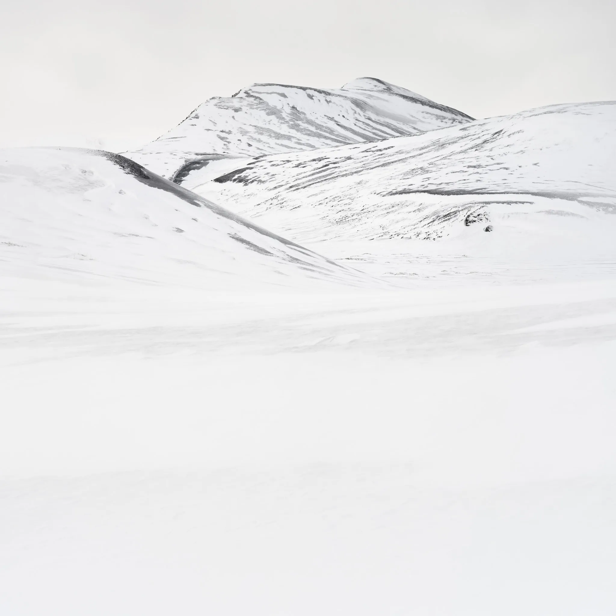 Layered snow-covered slopes with faint dark streaks rising to a distant ridge, nearly monochrome