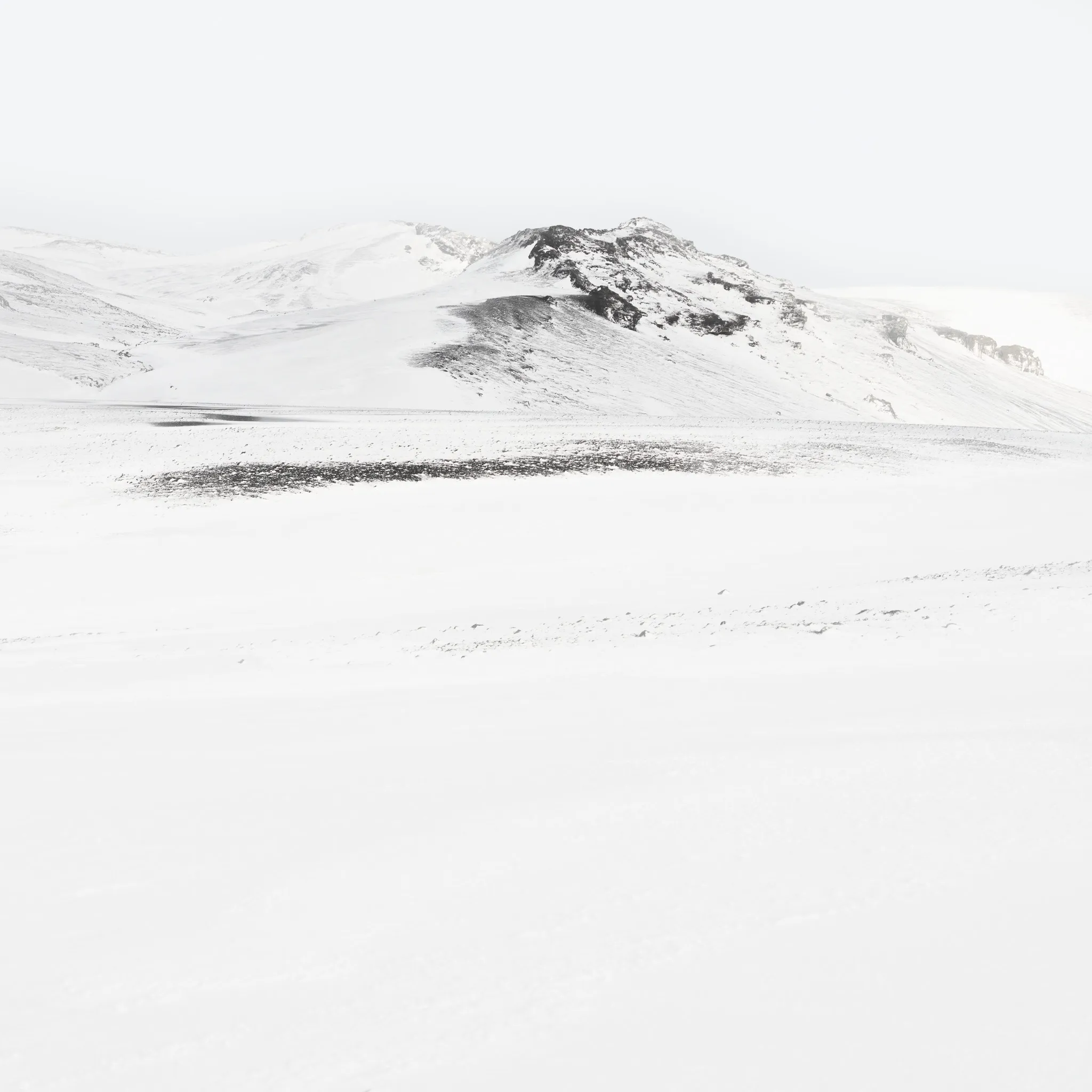 A snow mountain with a dark rocky flank above a white plain marked by a single dark streak