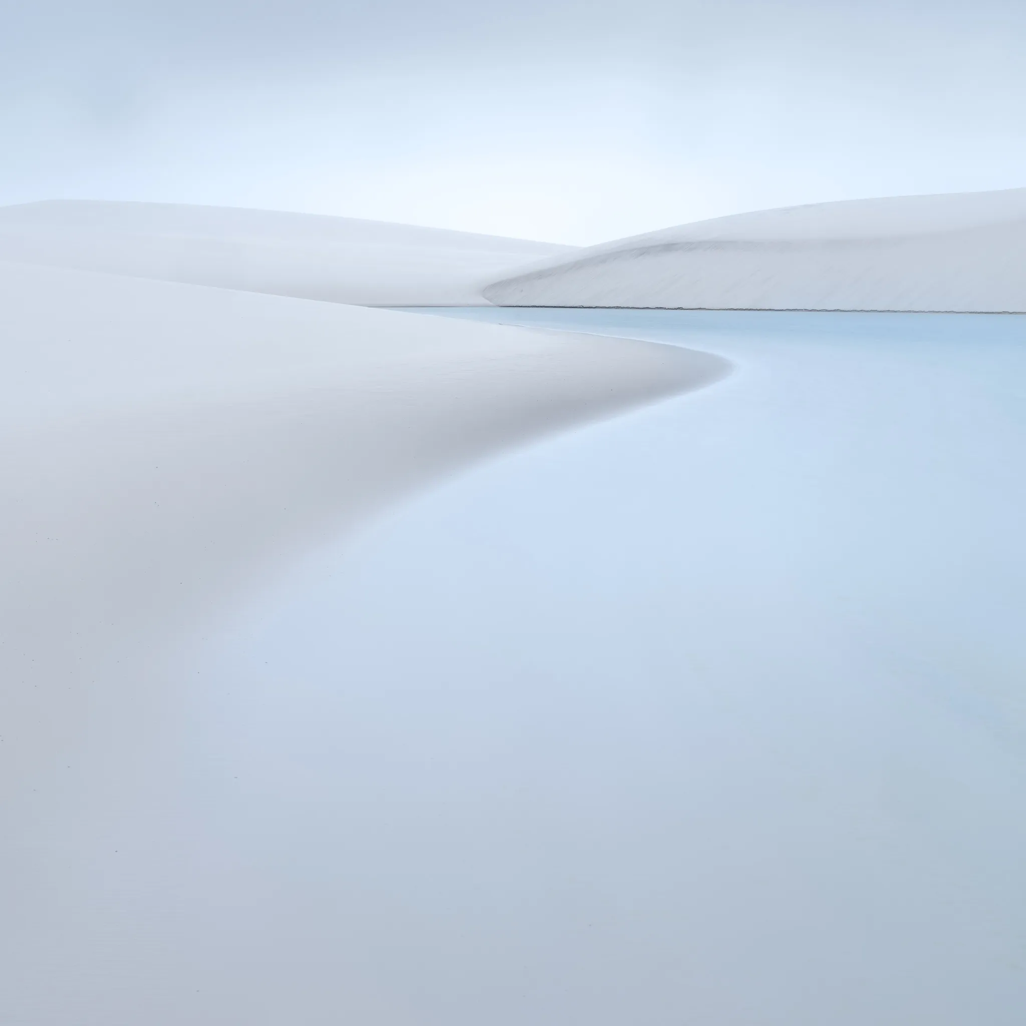A curving white dune edge meeting a pale blue lagoon, more dunes beyond