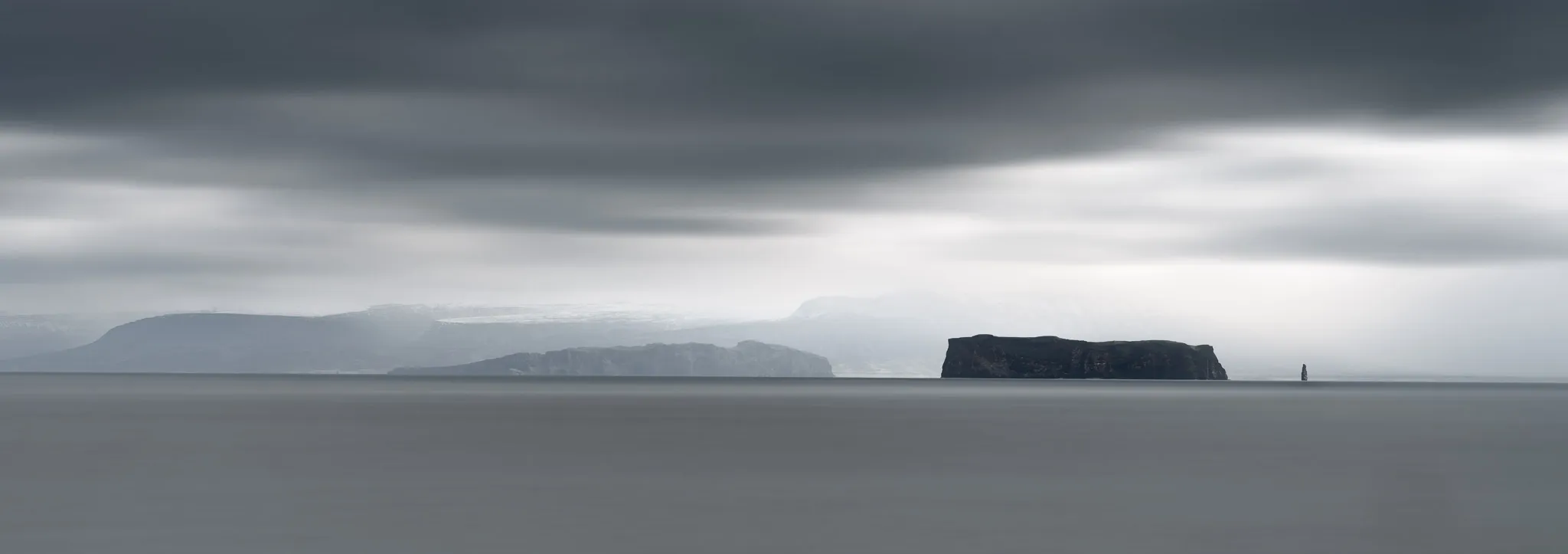 Flat-topped islands and a distant sea stack under a brooding sky above a silky, long-exposure sea