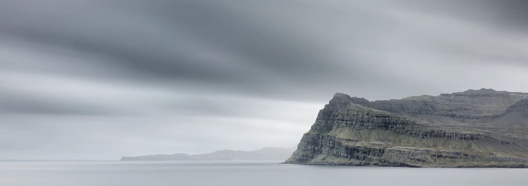 A stratified green-and-black sea cliff rising from calm, misty water under a brooding grey sky
