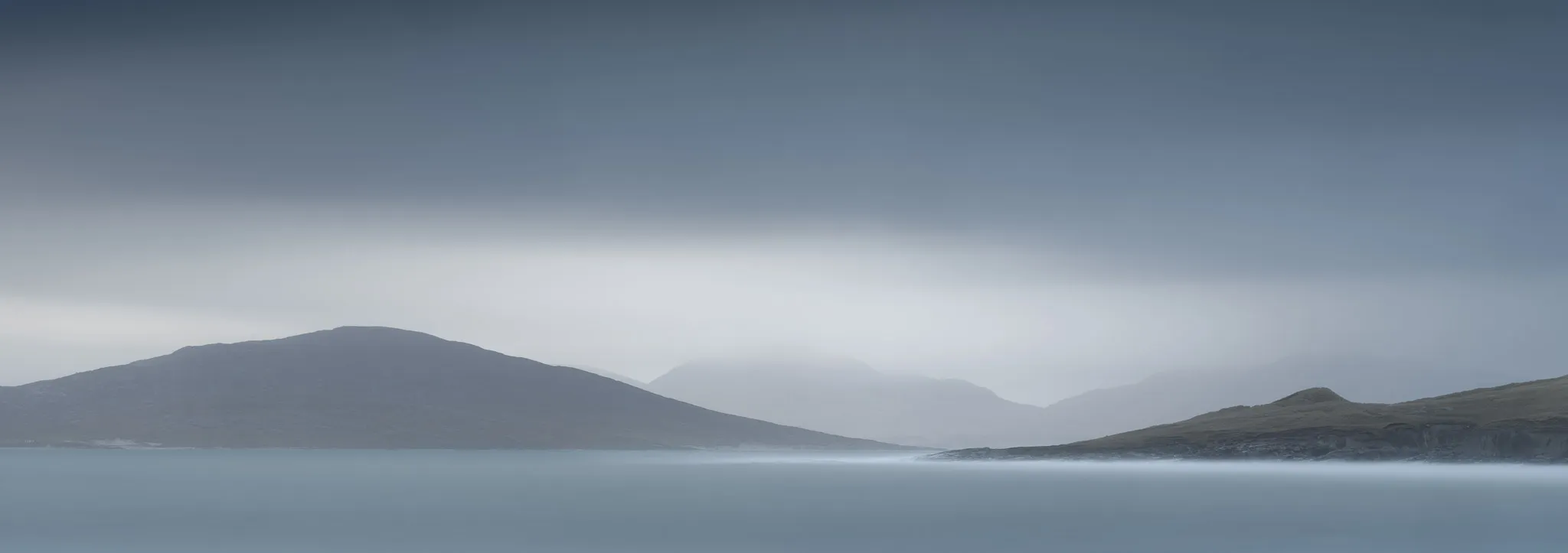 Layered misty hills above a smooth, pale turquoise sea in soft, silvery light