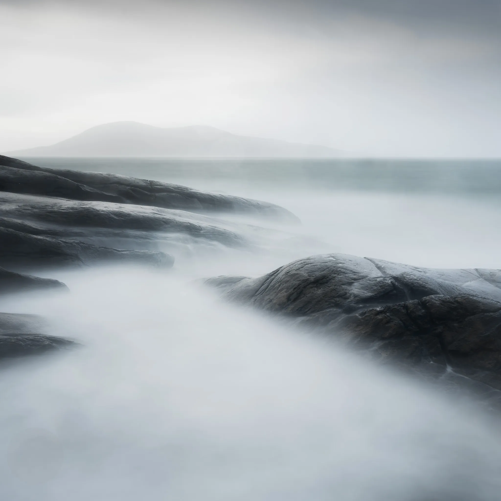 Misty long-exposure sea washing over dark coastal rocks, a headland fading into fog