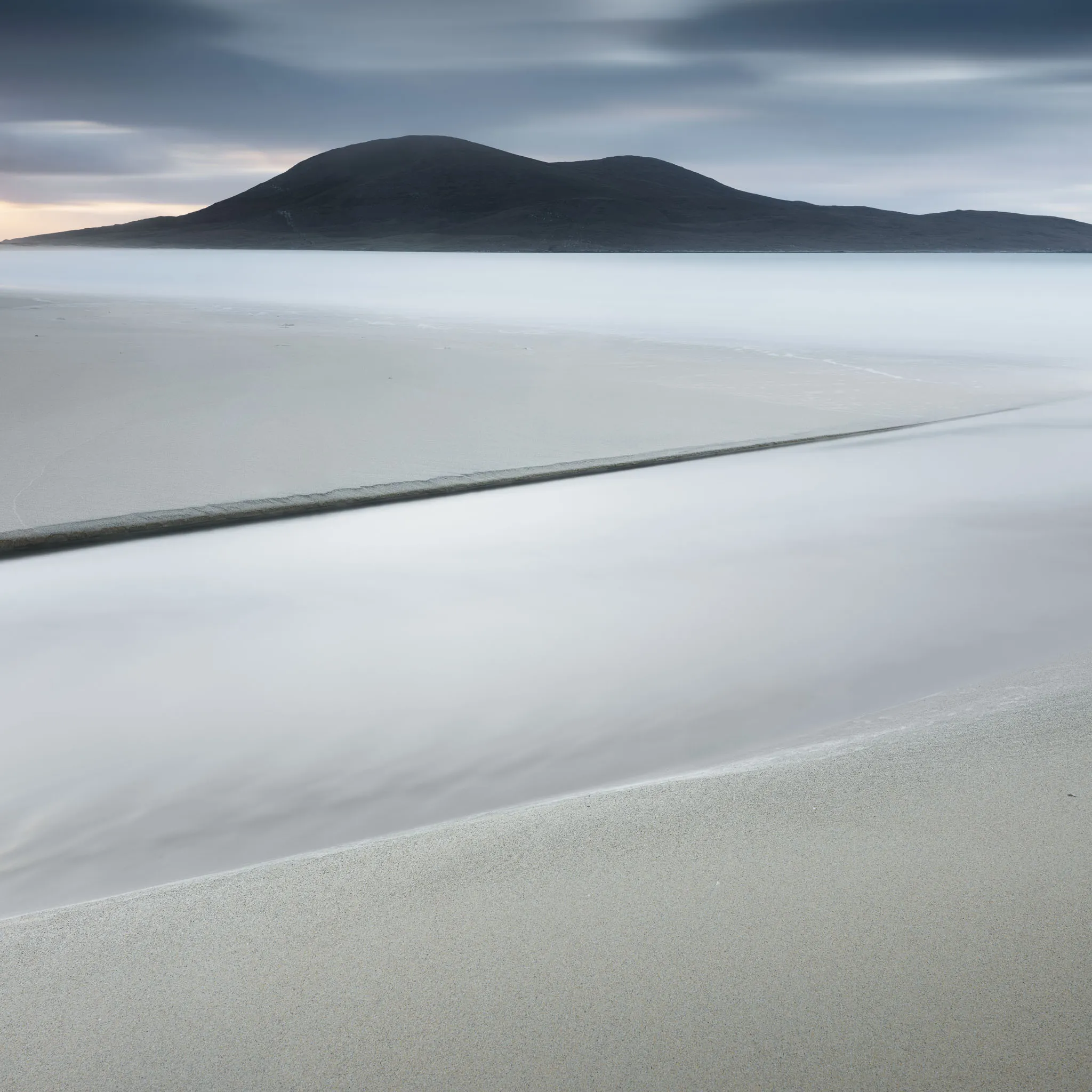 A dark hill above a beach with a bold diagonal sand line and silky water